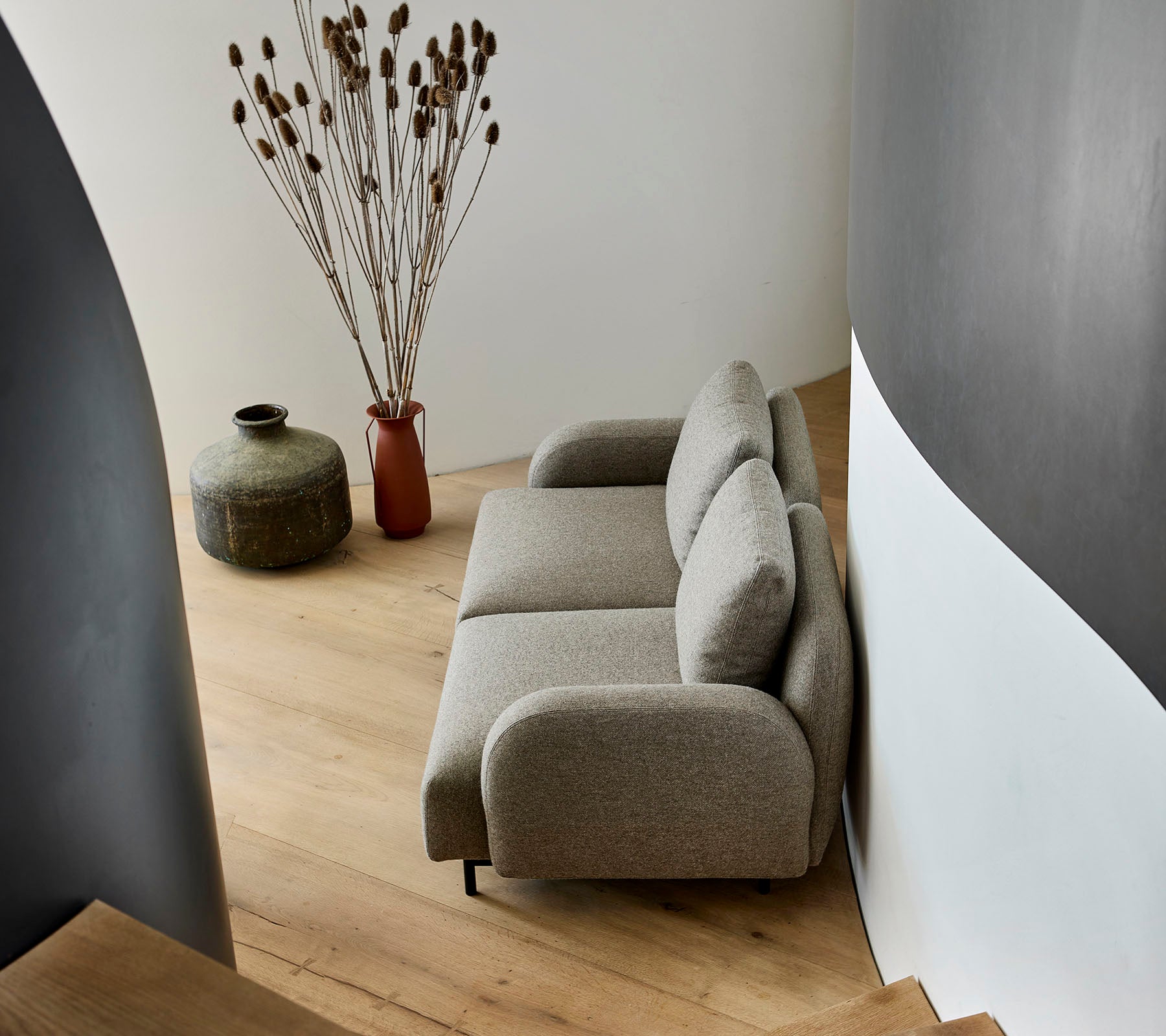 Een gezellige beige bank geplaatst naast een decoratieve vaas en gedroogde planten, tegen een gebogen muur en houten vloeren.