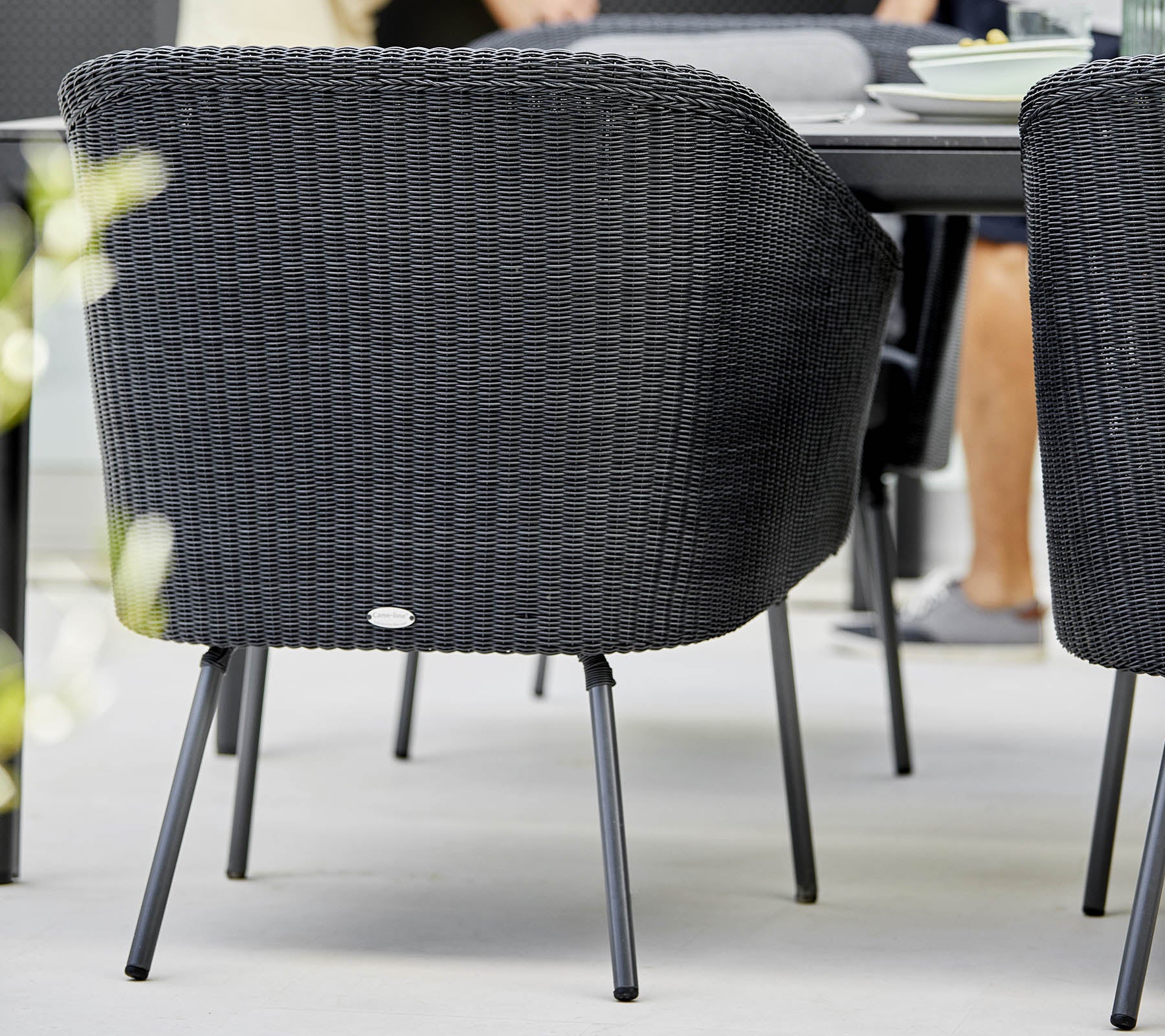 Zwarte gevlochten stoel met gebogen rugleuning en metalen poten, geplaatst bij een eettafel met zachte focus op de omringende elementen.