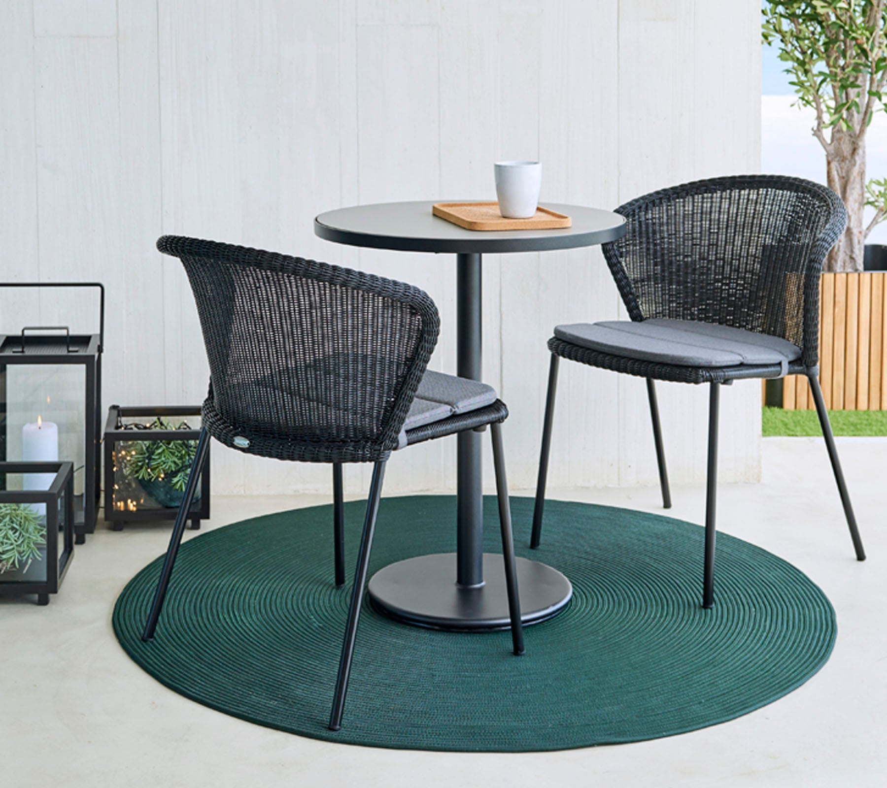 Een ronde tafel met twee zwarte gevlochten stoelen op een groen tapijt, vergezeld van een koffiekop en een houten plank. Decoratieve lantaarns op de achtergrond.