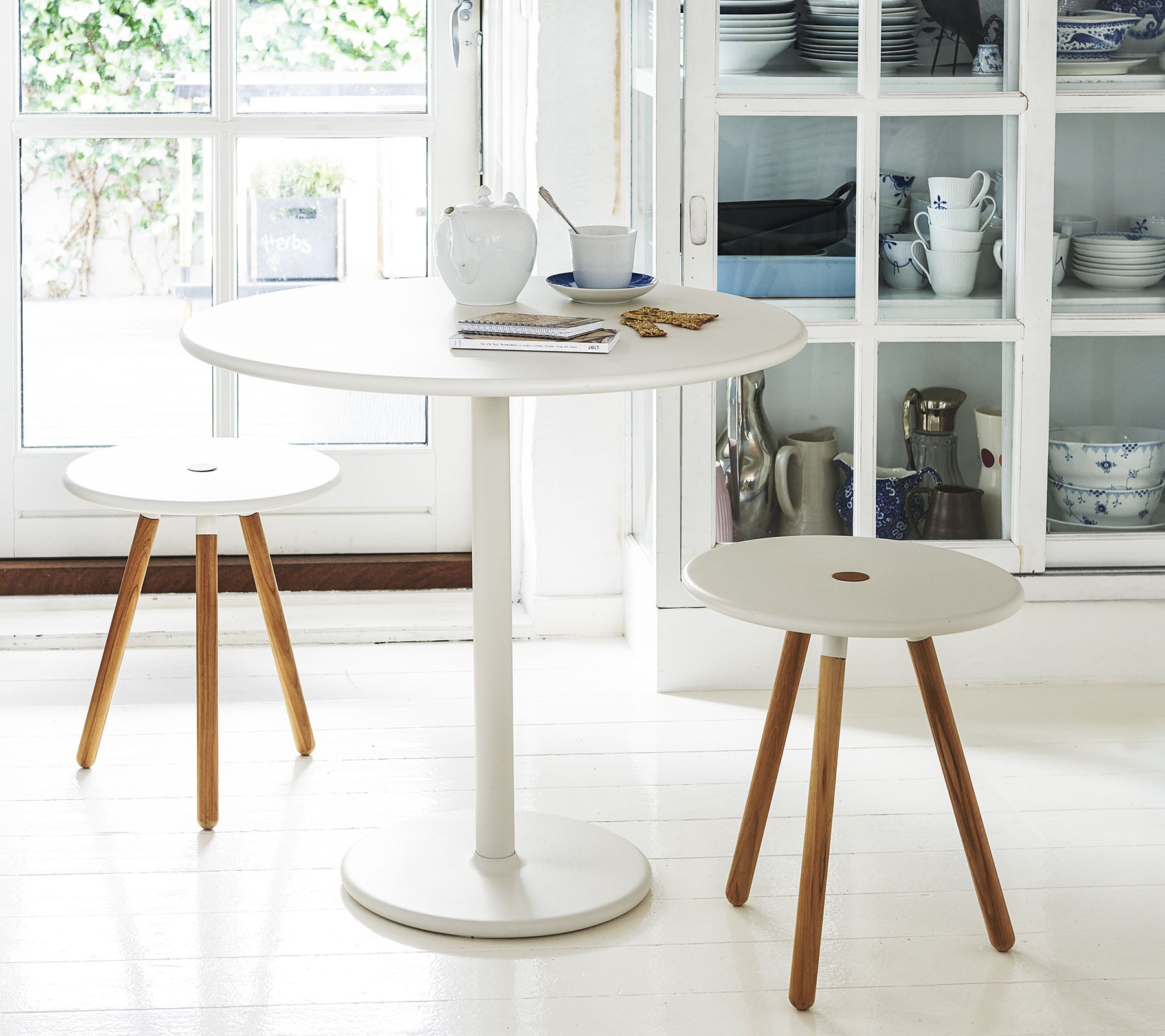 Een ronde witte tafel vergezeld van twee minimalistische krukken met houten poten, voor een achtergrond van planken met keukengerei.