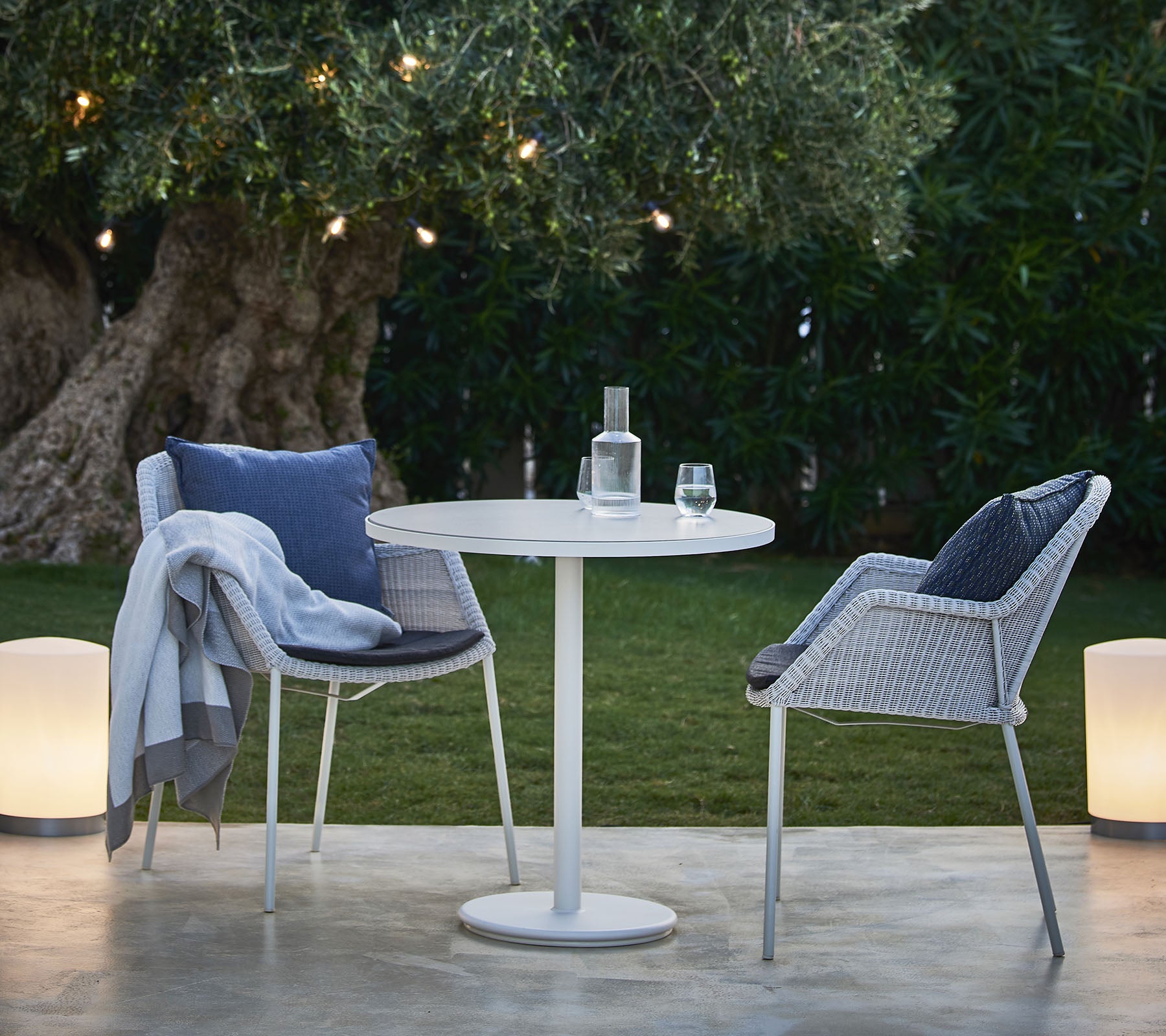 Een kleine ronde tafel met twee stijlvolle stoelen, een fles water en een glas, geplaatst temidden van weelderig groen en zacht gloeiende lichten.