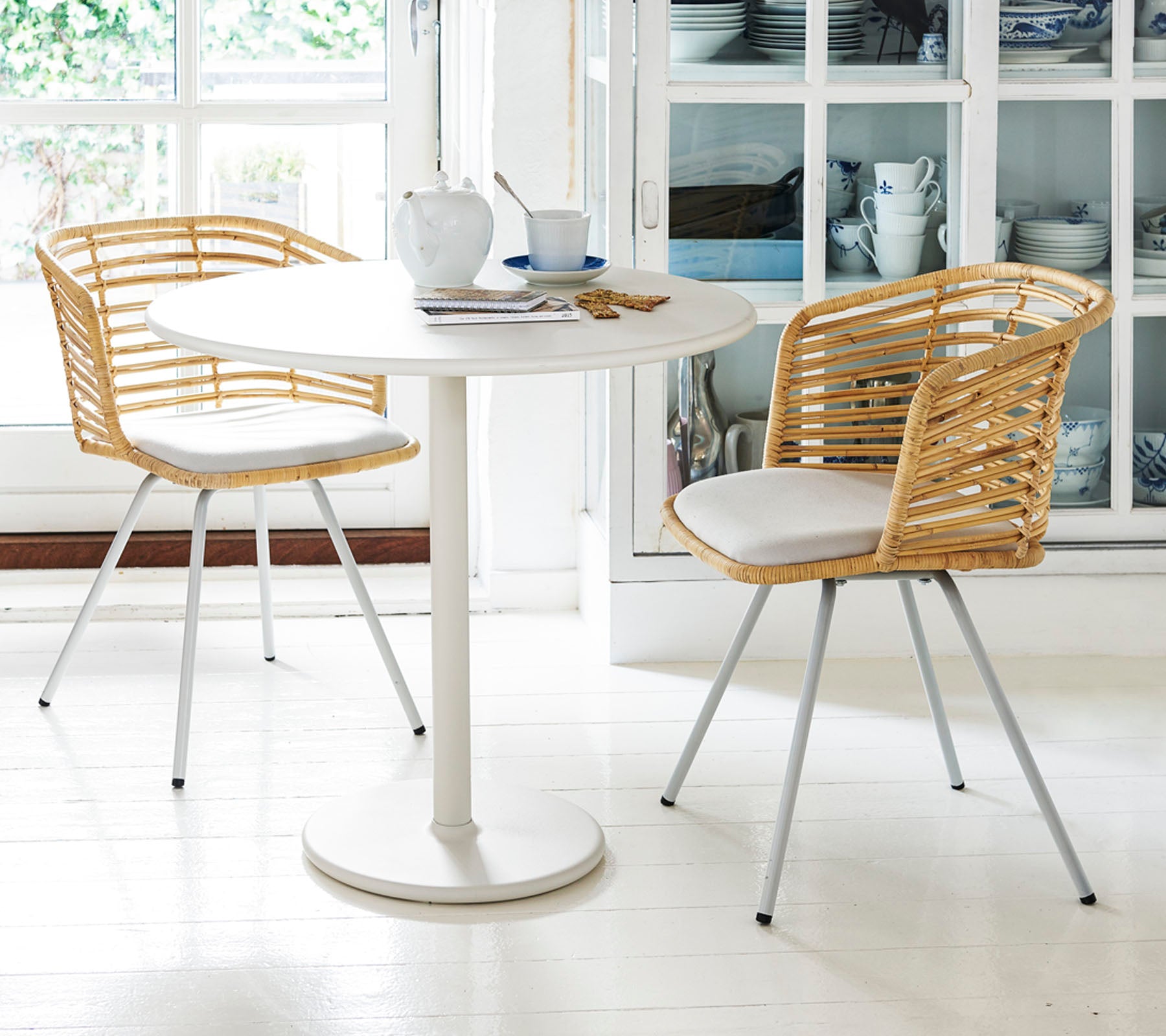 Een ronde tafel met twee stijlvolle stoelen heeft gevlochten ruggen en kussens, gedekt met theekopjes en een schoteltje, waarmee een gezellige eetruimte wordt gecreëerd.