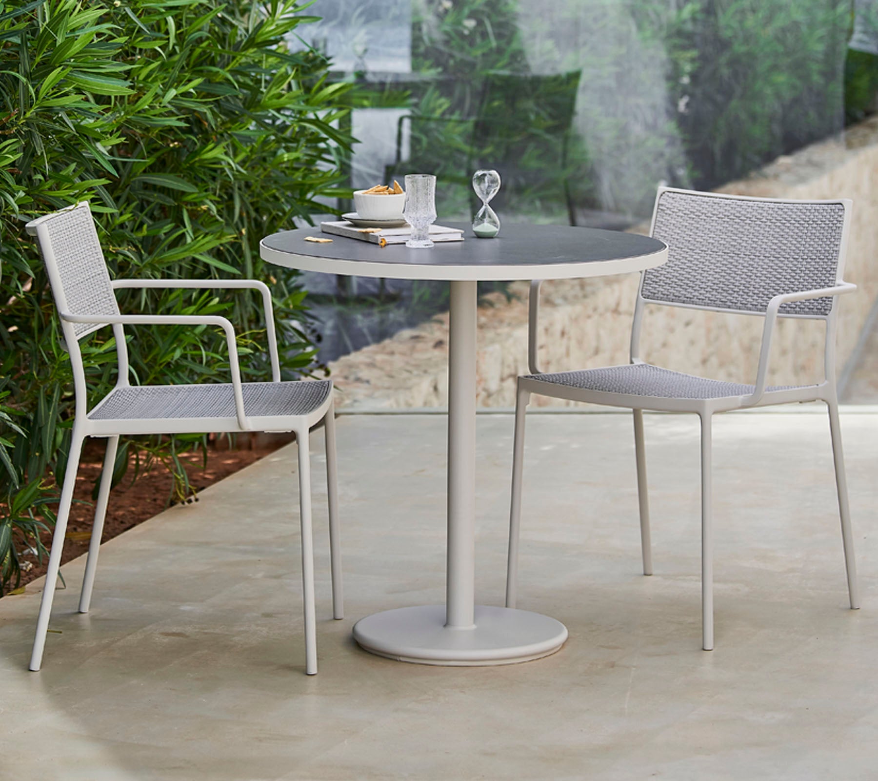 Een stijlvol ronde tafel omringd door twee moderne stoelen, gedekt met servies en een decoratief item, omringd door weelderig groen.