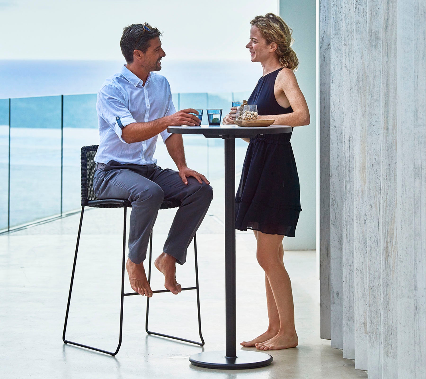 Een man en vrouw genieten van drankjes aan een hoge tafel, in gesprek, met een pittoreske achtergrond van water in de verte.