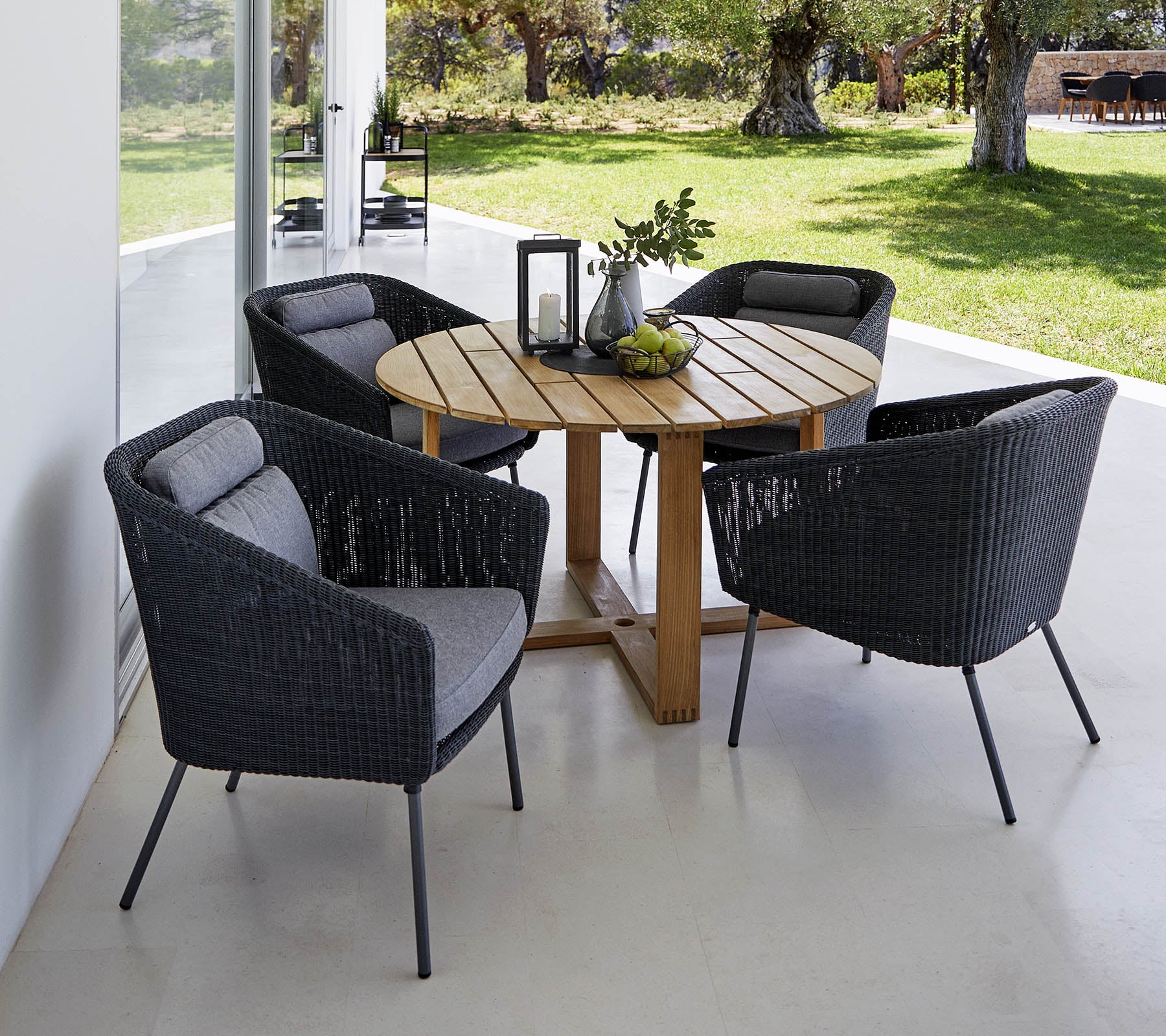 Een ronde houten tafel omringd door vier stijlvolle zwarte gevlochten stoelen met grijze kussens, geaccentueerd door een decoratieve lantaarn en groen.