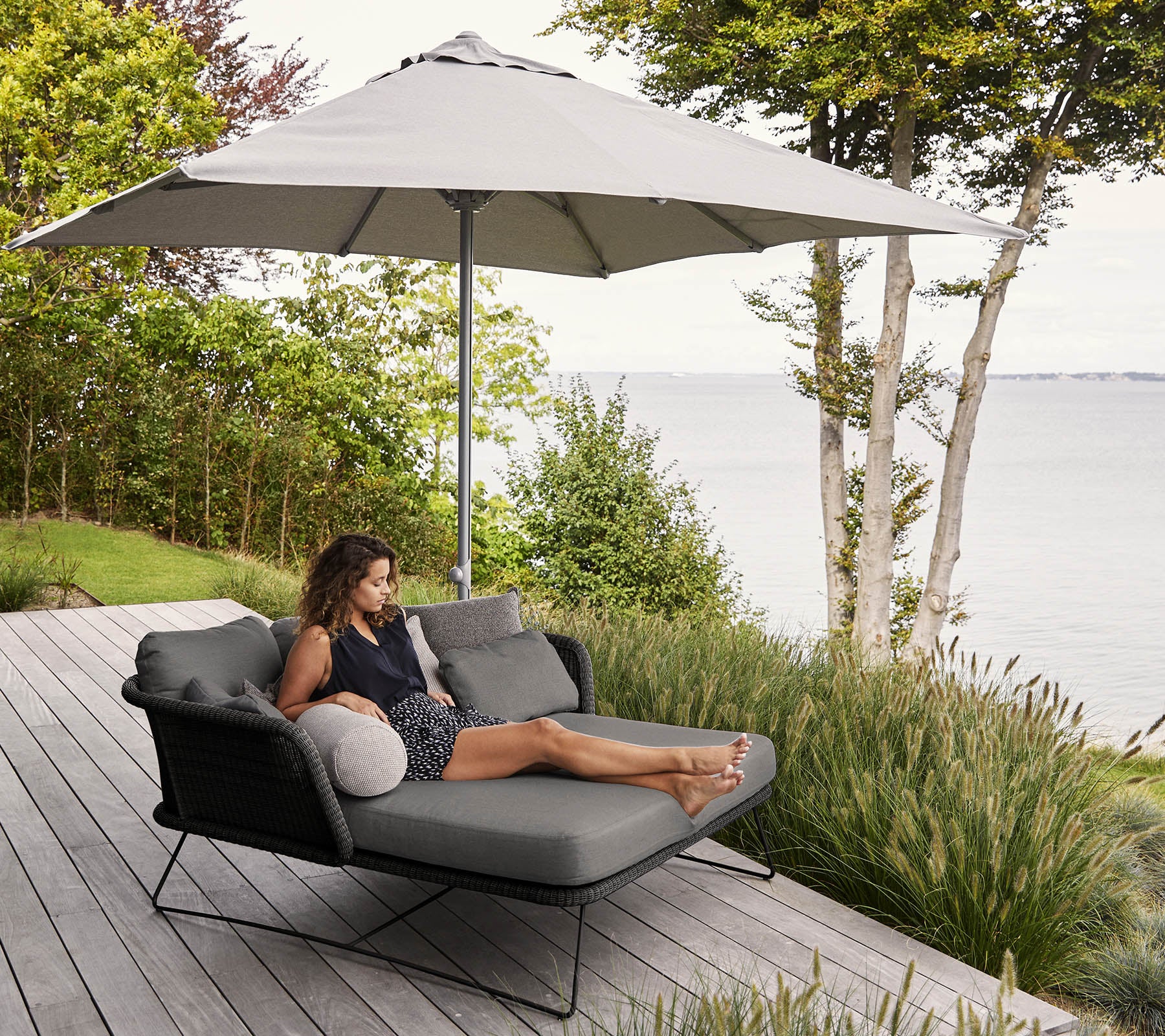 Een vrouw ontspant op een moderne loungechair met een grote parasol, omringd door groen en met uitzicht op het water.