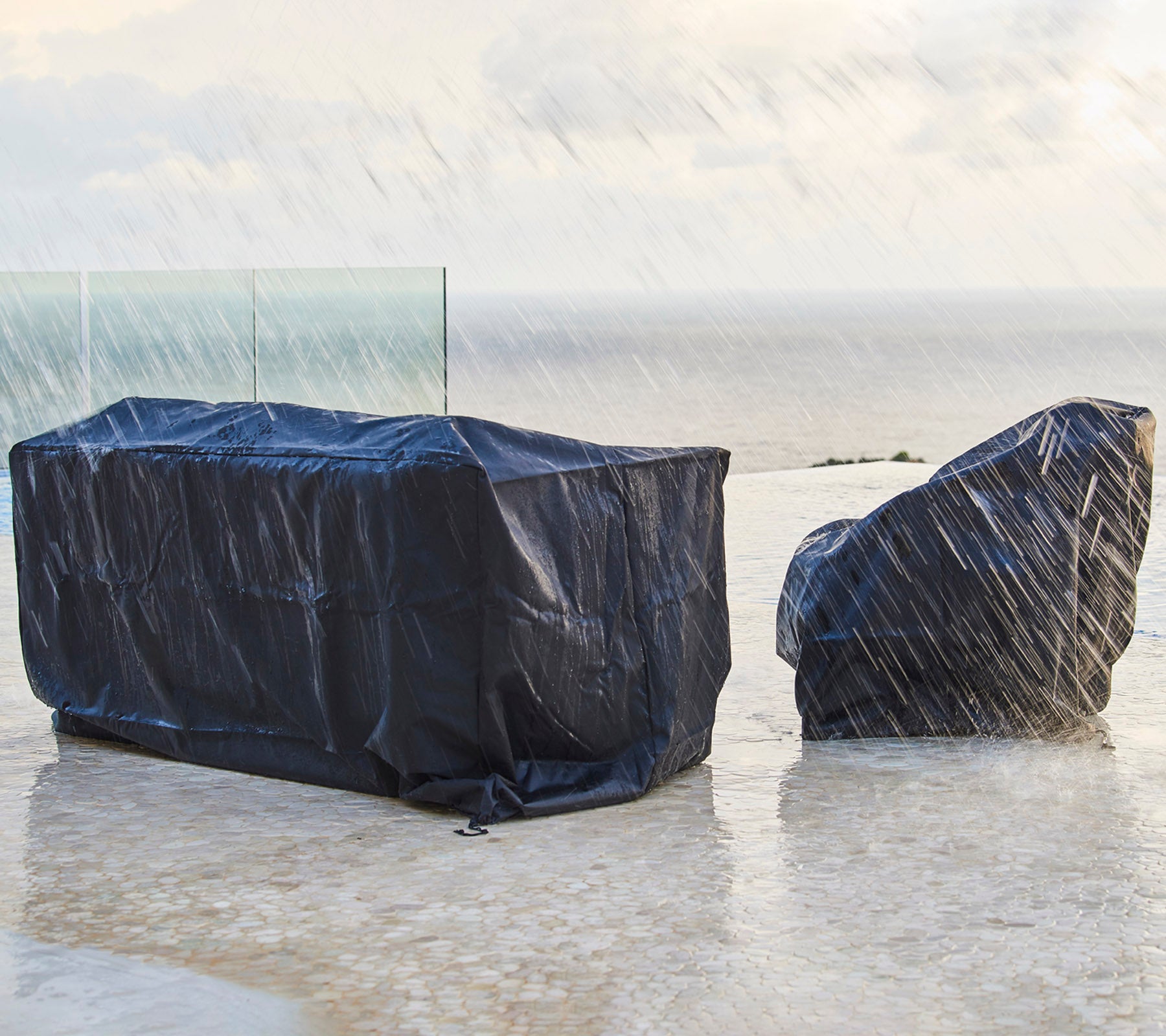 Tuinfurnitur bedekt met donkere beschermhoezen, beschermd tegen regen, met een achtergrond van een bewolkte lucht en de oceaan.