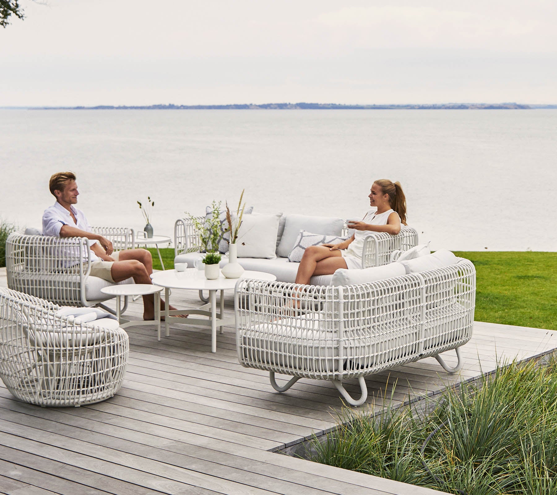 Twee personen ontspannen in een stijlvolle, comfortabele zitruimte met witte wicker meubels en een salontafel, met uitzicht op een pittoresk waterlandschap.