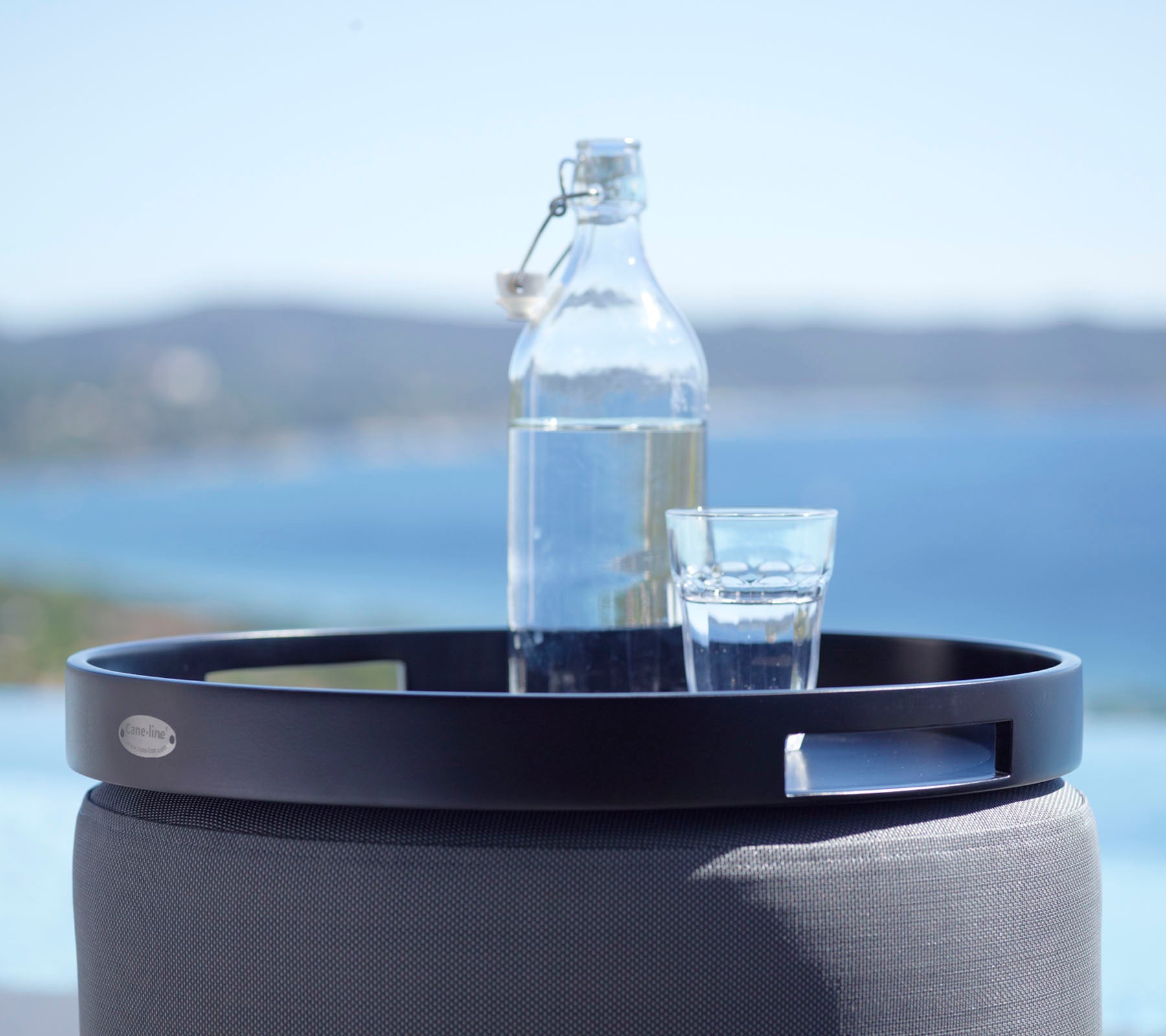 Een zwart rond dienblad houdt een glas water en een glazen fles, tegen een schilderachtige achtergrond van de zee en verre heuvels.