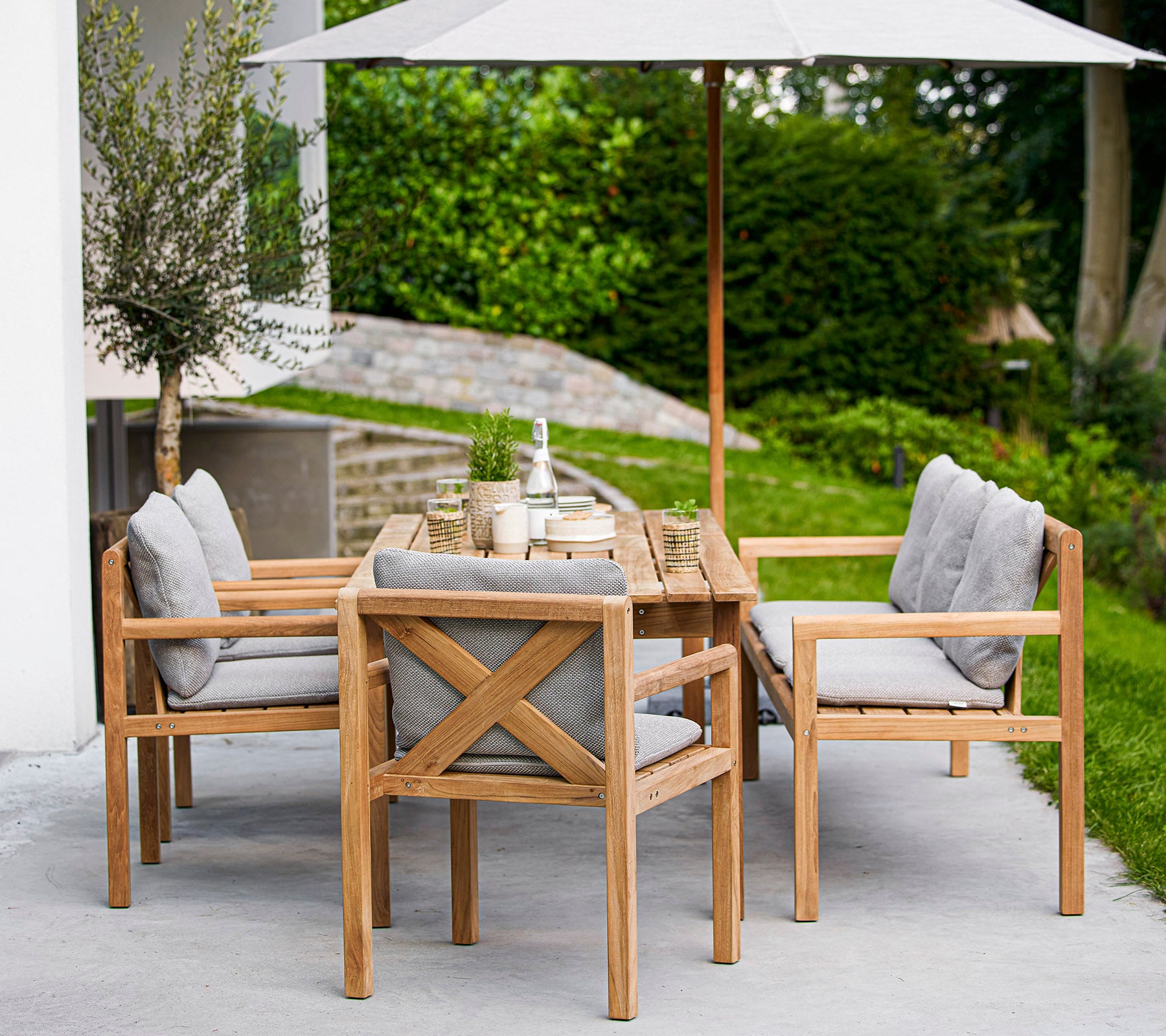 Houten eetset met een rechthoekige tafel en gestoffeerde stoelen, met een grote parasol die schaduw biedt. Groen is zichtbaar op de achtergrond.