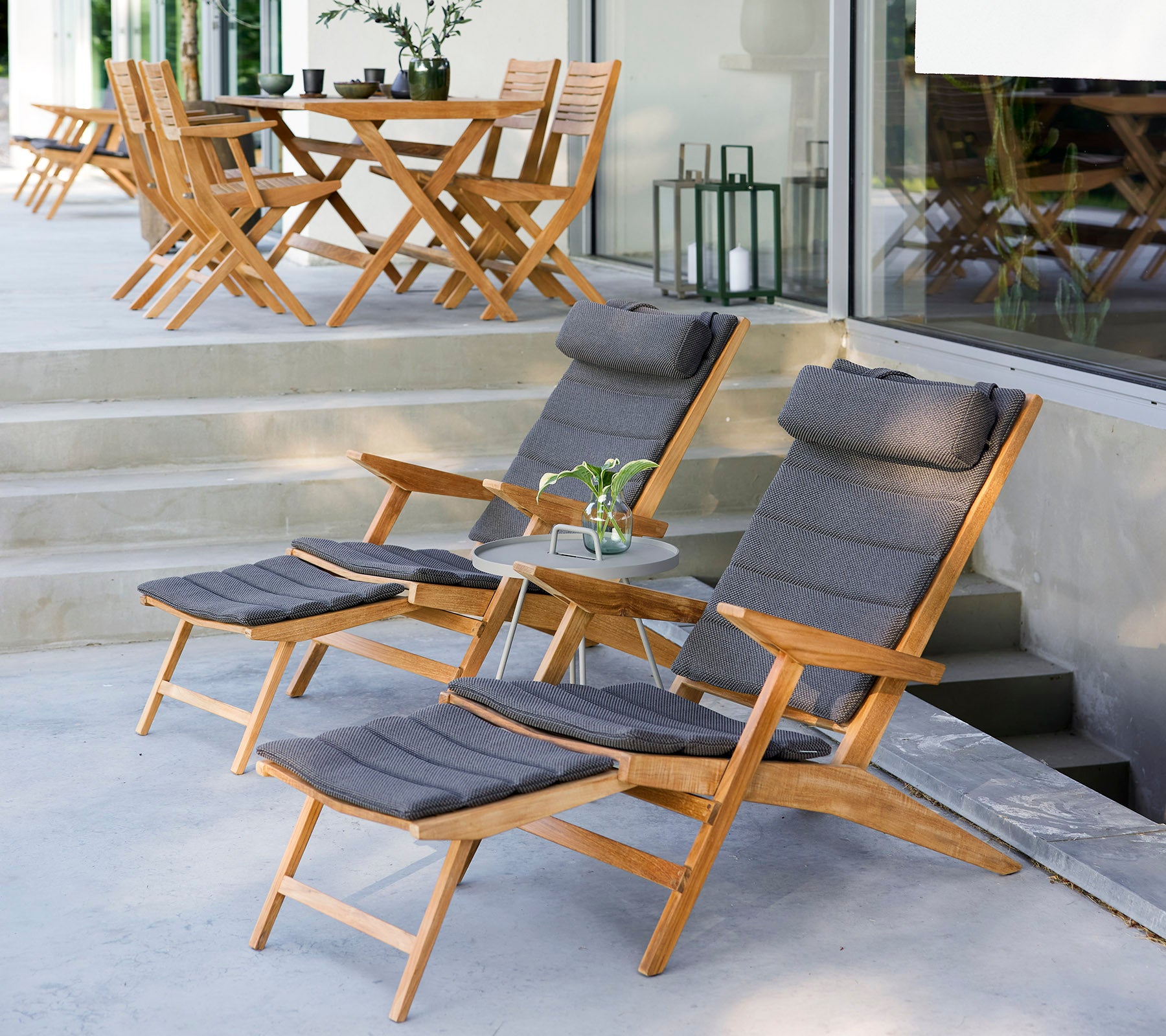Twee houten loungestoelen met kussens, een kleine bijzettafel en groen in een stijlvolle buitensetting met extra stoelen op de achtergrond.
