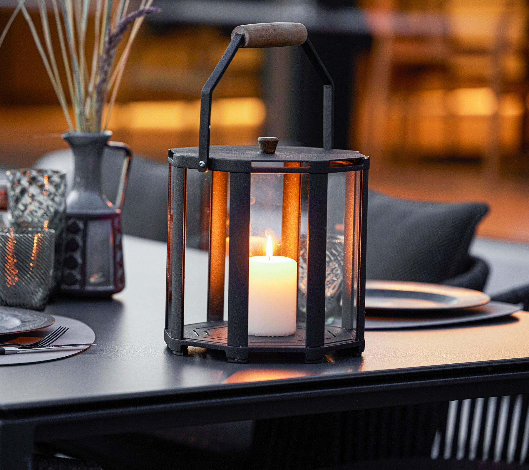 Een stijlvolle lantaarn met een kaars erin, met een metalen frame en houten handvat, geplaatst op een tafel met decoratieve elementen eromheen.