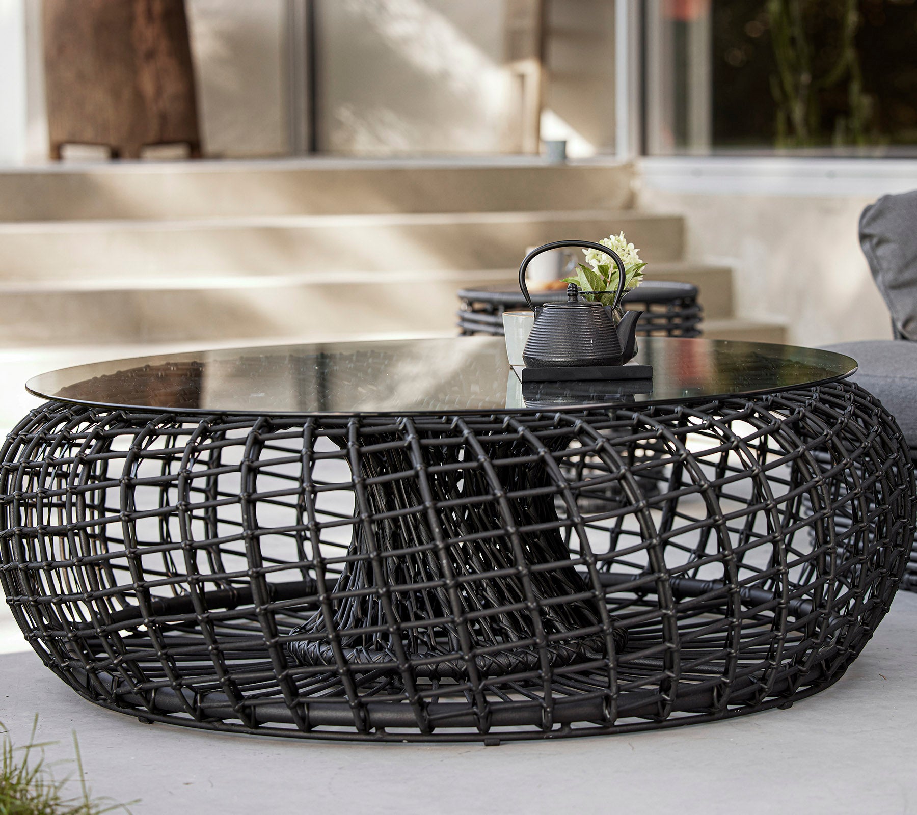Een modern, rond salontafel met een geweven zwarte basis en een glazen blad, met decoratieve items zoals een plant en een kleine kom.