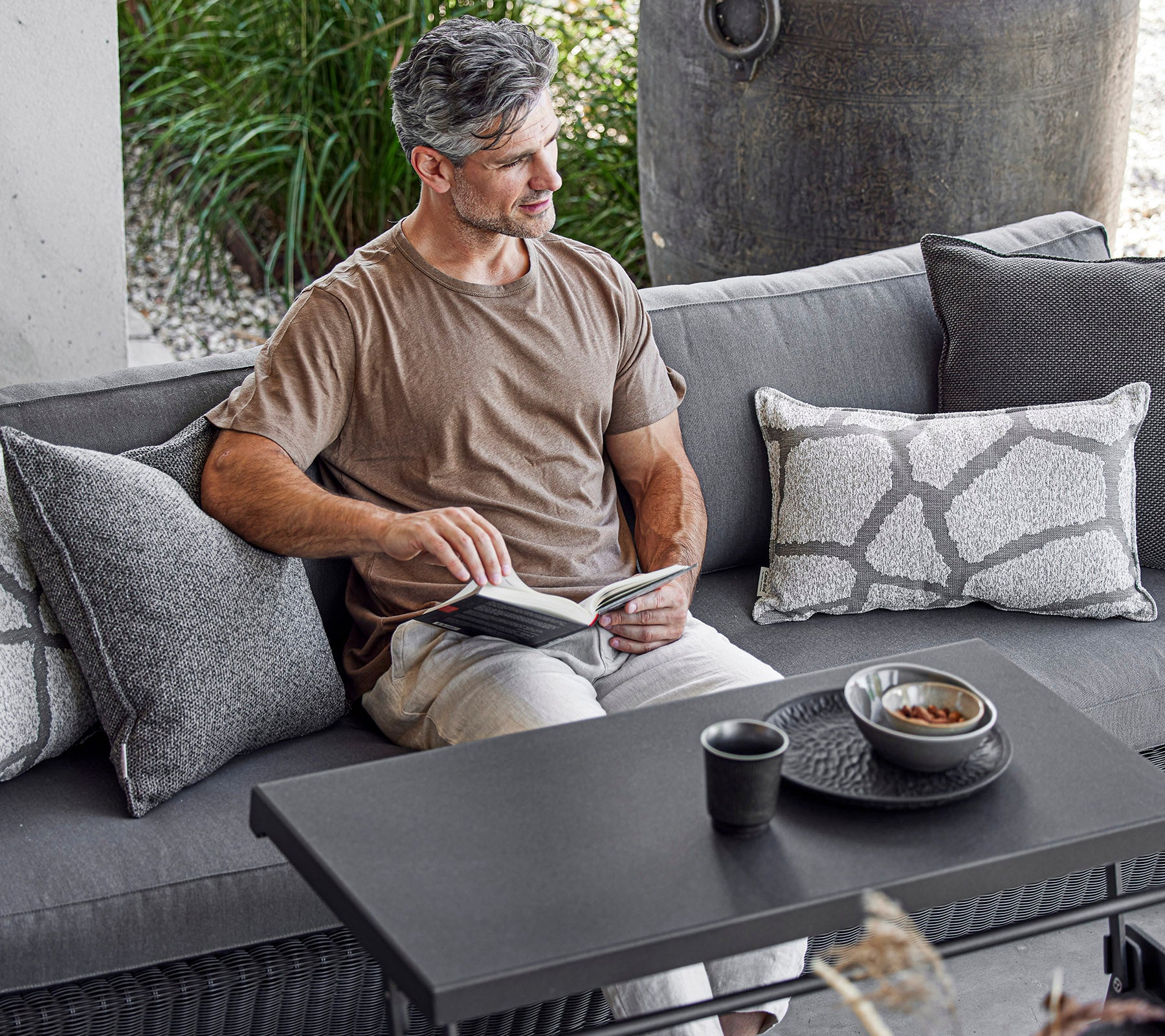 Een man zit comfortabel op een bank, leest een boek met een kop en een kom op een salontafel naast hem, omringd door decoratieve kussens.