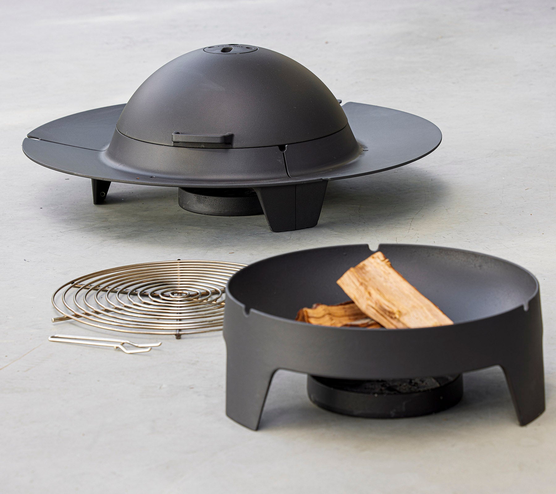 Een moderne vuurkorfsset met een strak, rond ontwerp met een koepeldeksel, vergezeld van een grillrek en houtstukken.