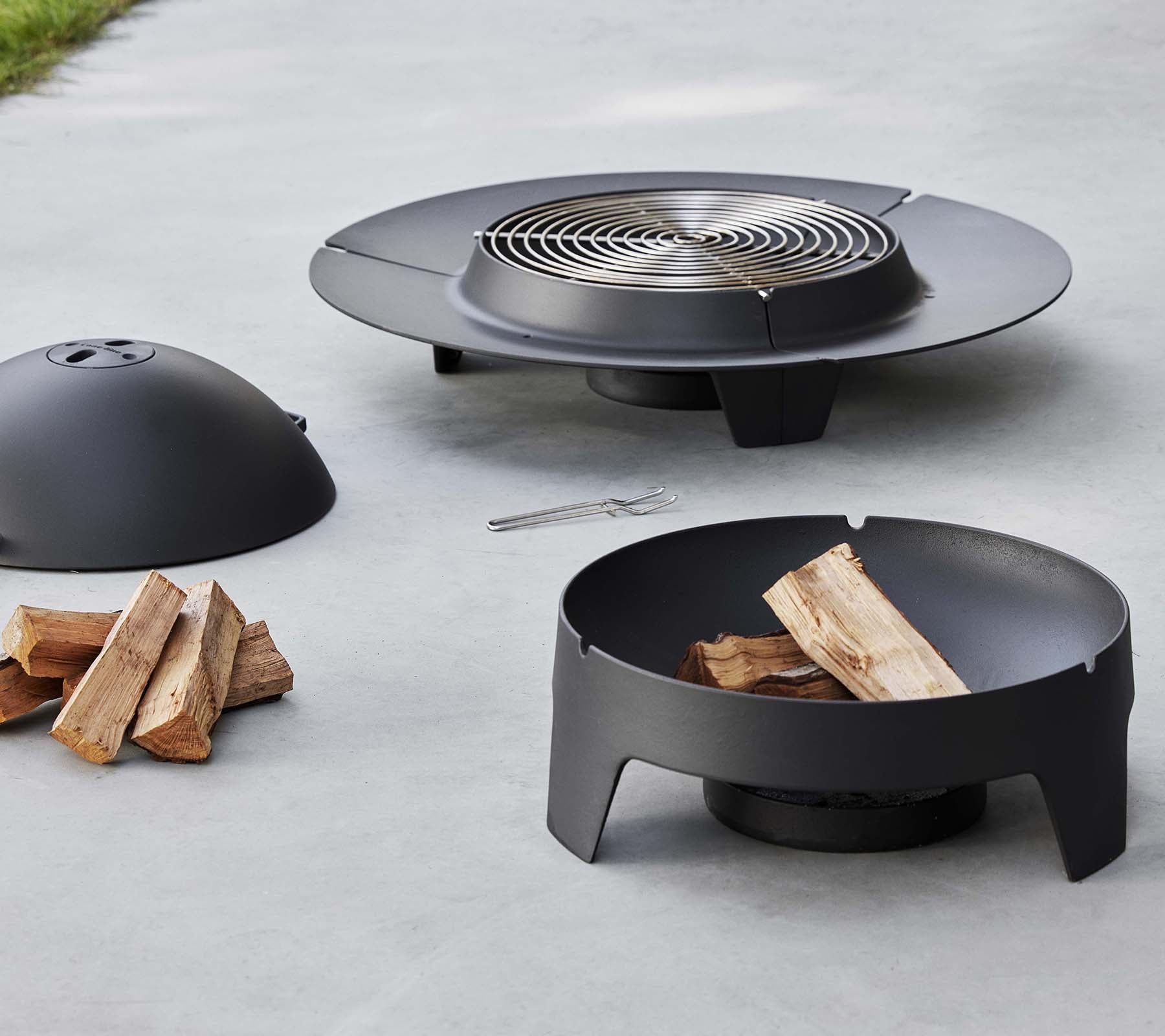 Een slanke zwarte vuurplaats met een grillrooster, een beschermhoes en een basis, vergezeld van gestapeld hout.