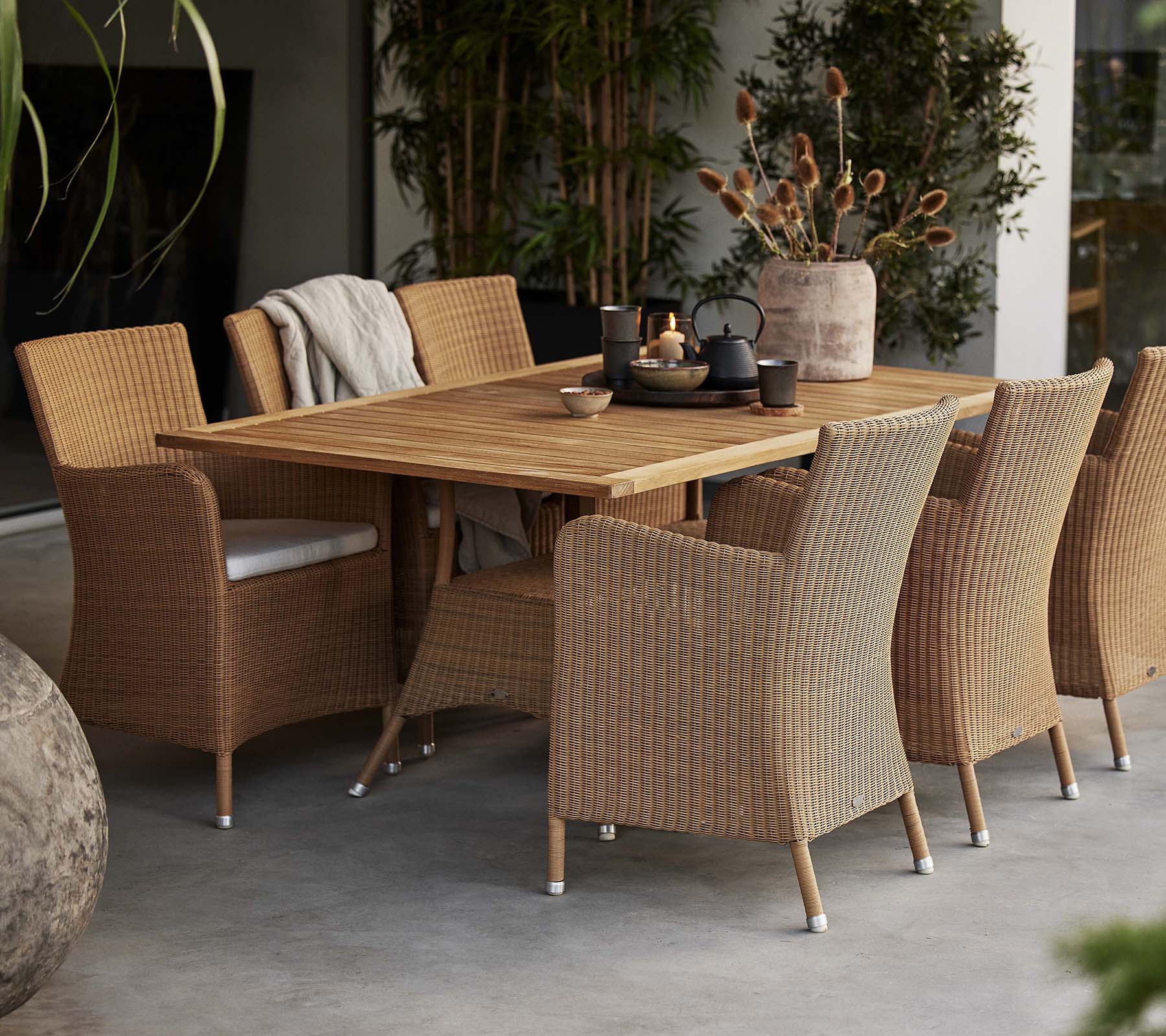 Een eetopstelling met een rustieke houten tafel omringd door geweven Hampsted-stoelen, aangevuld met decoratieve planten.