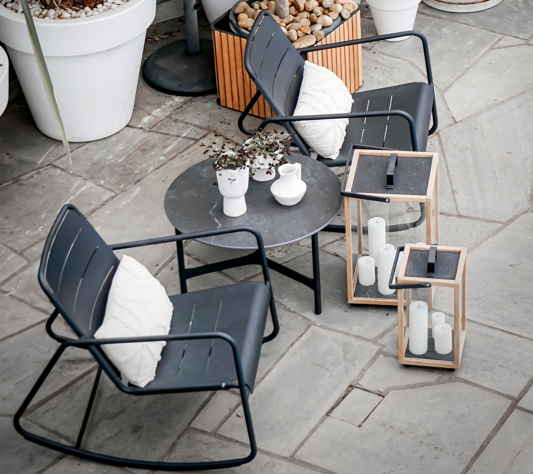Twee moderne schommelstoelen en een ronde tafel met decoratieve planten en lantaarns, geplaatst op een gestructureerd stenen oppervlak.