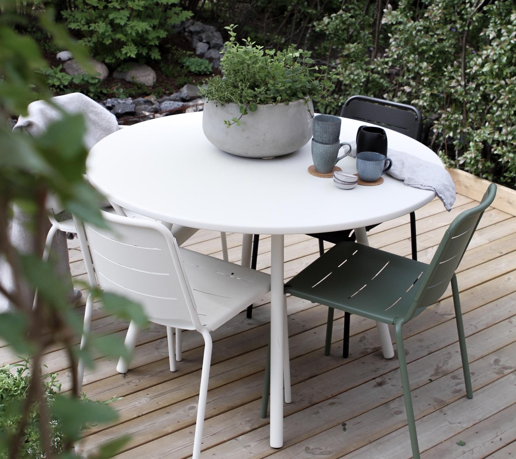 Een ronde tafel omringd door vier stoelen, met een potplant en koffiemokken op het tafelblad. Lush groen in de achtergrond.