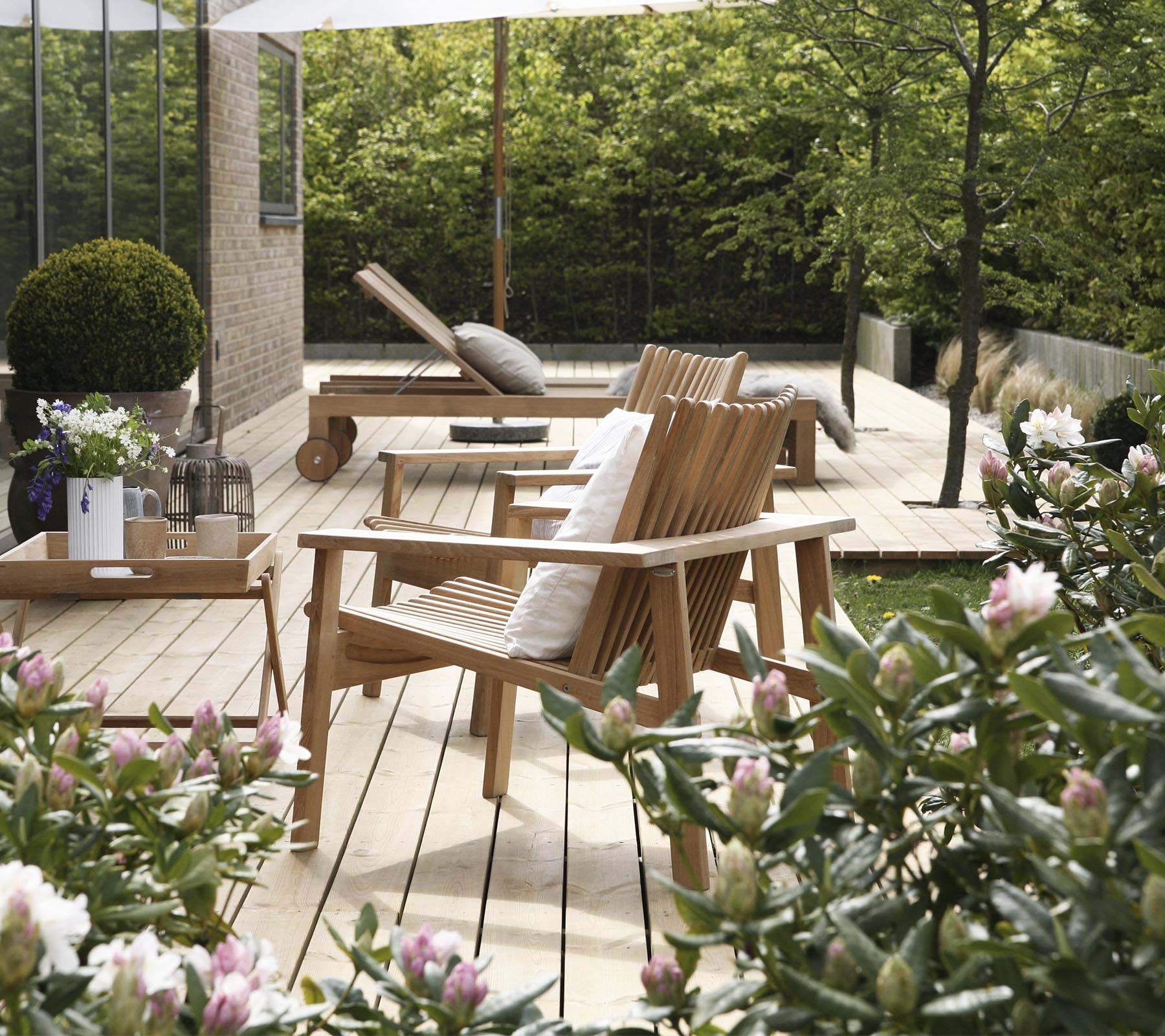 Houten stoelen en een tafel zijn omringd door groen en bloemen, wat een serene en uitnodigende buitenzitplaats creëert.