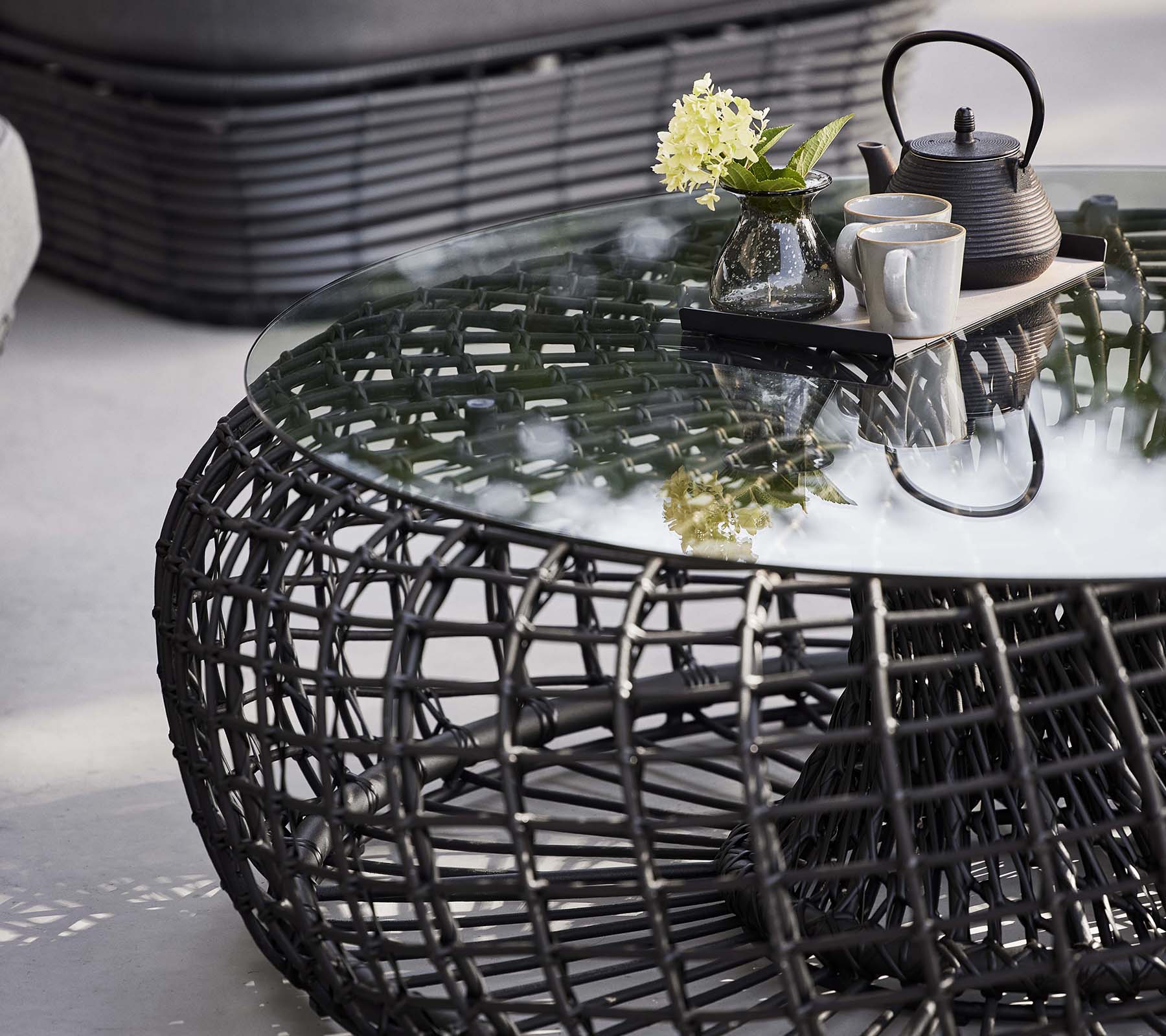 Nest koffie tafel met een gevlochten basis, een glazen blad en een theeset met bloemen voor een stijlvolle, moderne uitstraling.