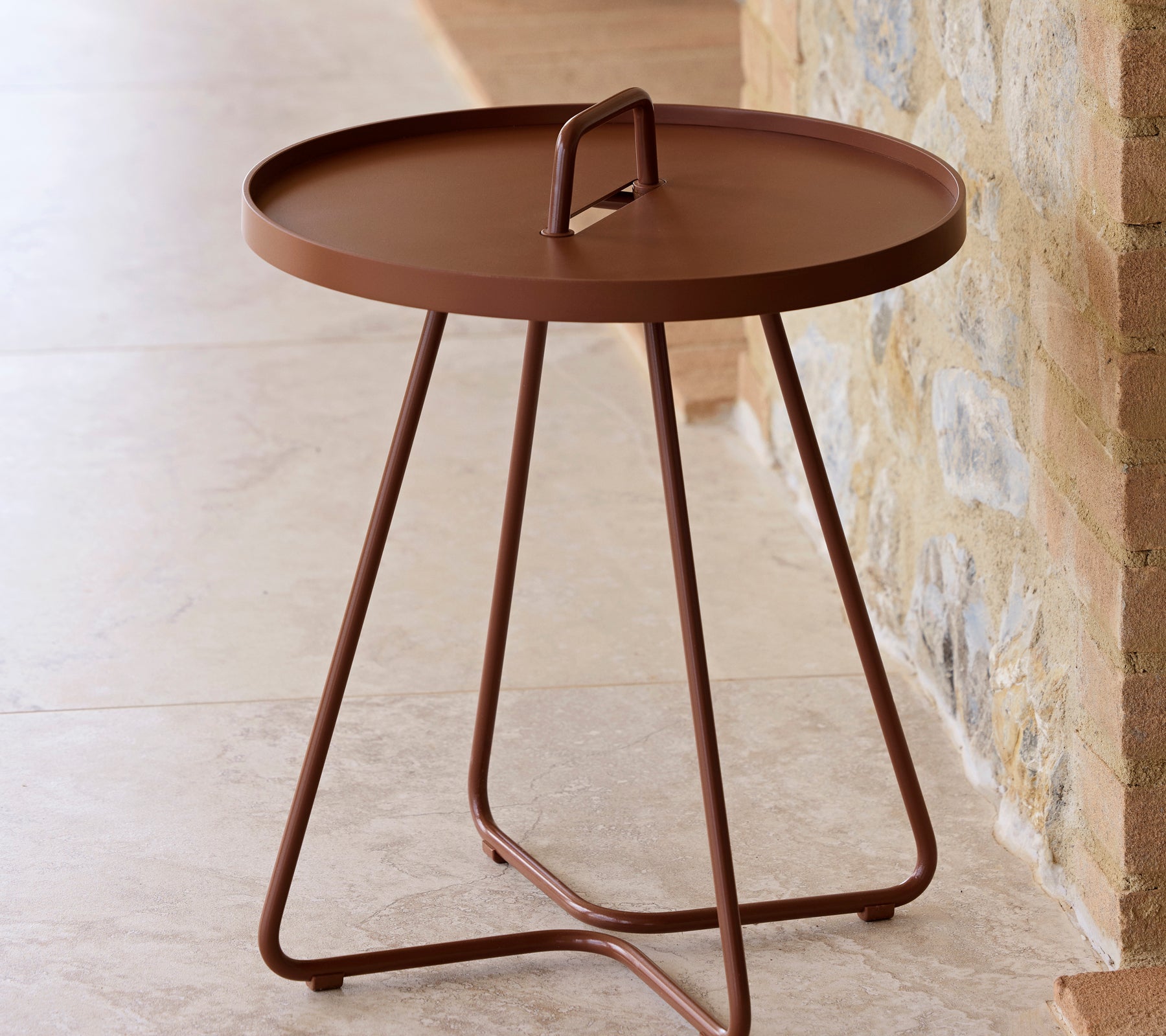 Een ronde tafel in een woestijnrode afwerking met een handgreep en drie slanke poten, ontworpen voor veelzijdigheid en gemakkelijke verplaatsing.