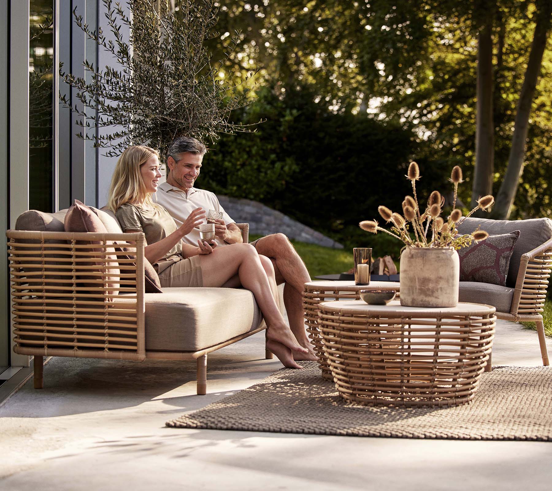 Een stel geniet van drankjes op een Sense-sofa en lounge stoel, vergezeld van een stijlvolle mand koffie tafel en een decoratieve vaas.