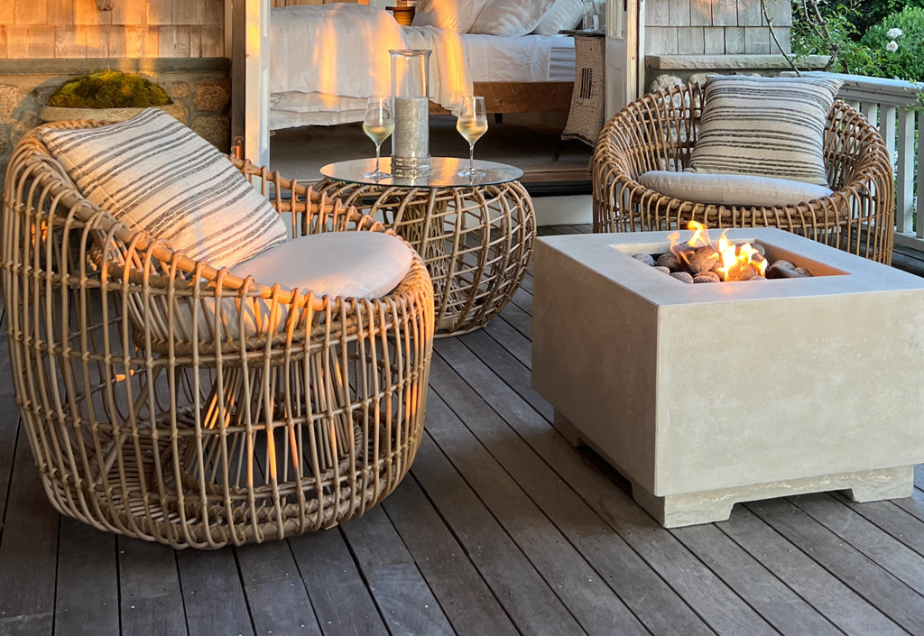 Two round lounge chairs with a round side table and fire pit table on a veranda at sunset