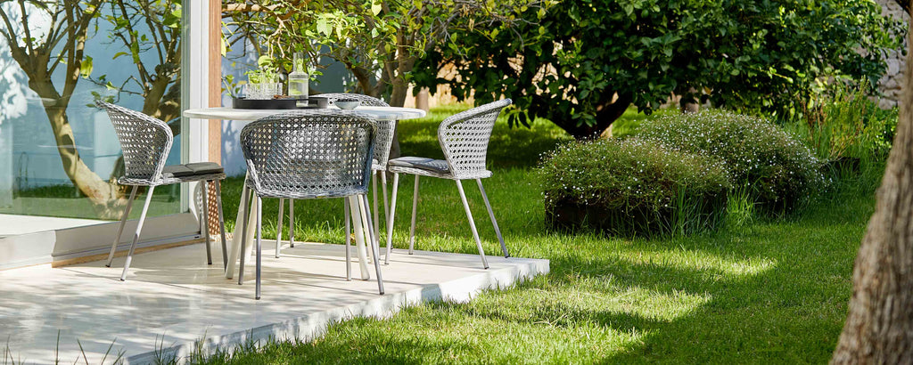 A modern table set with four stylish chairs on a grassy area, surrounded by lush greenery and soft landscaping.