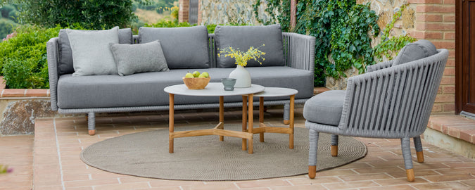 Stijlvolle grijze tuinsofa's met kussens, vergezeld van een ronde salontafel en een vaas met bloemen, geplaatst in een weelderige groene omgeving.