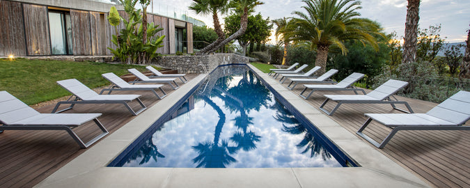 Een serene zwembadscène met een rij witte ligbedden naast rustig water, omgeven door weelderig groen en palmbomen.