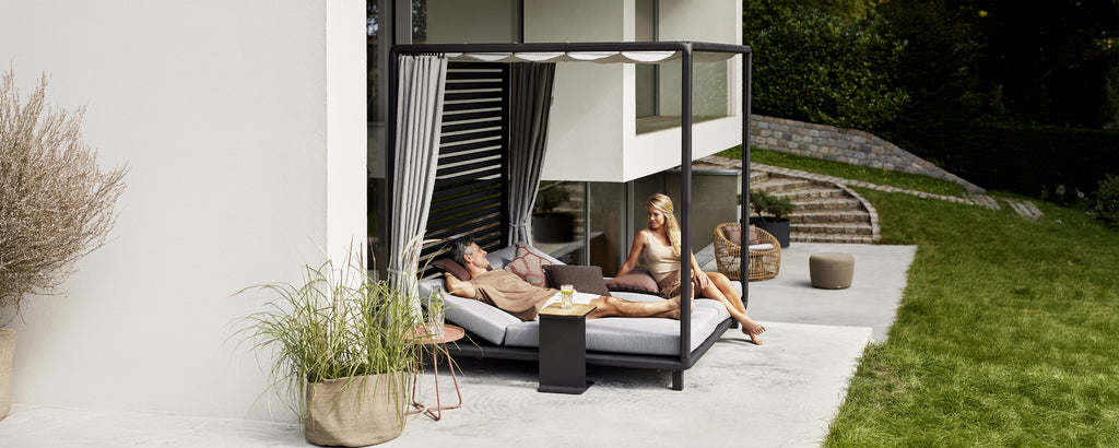 Moderne buitenlounge met een zwart daybed, grijze kussens en witte gordijnen.