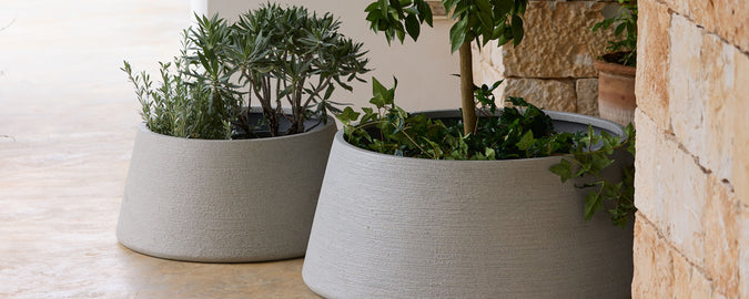 Twee moderne grijze plantenbakken met groen in een buitenomgeving.