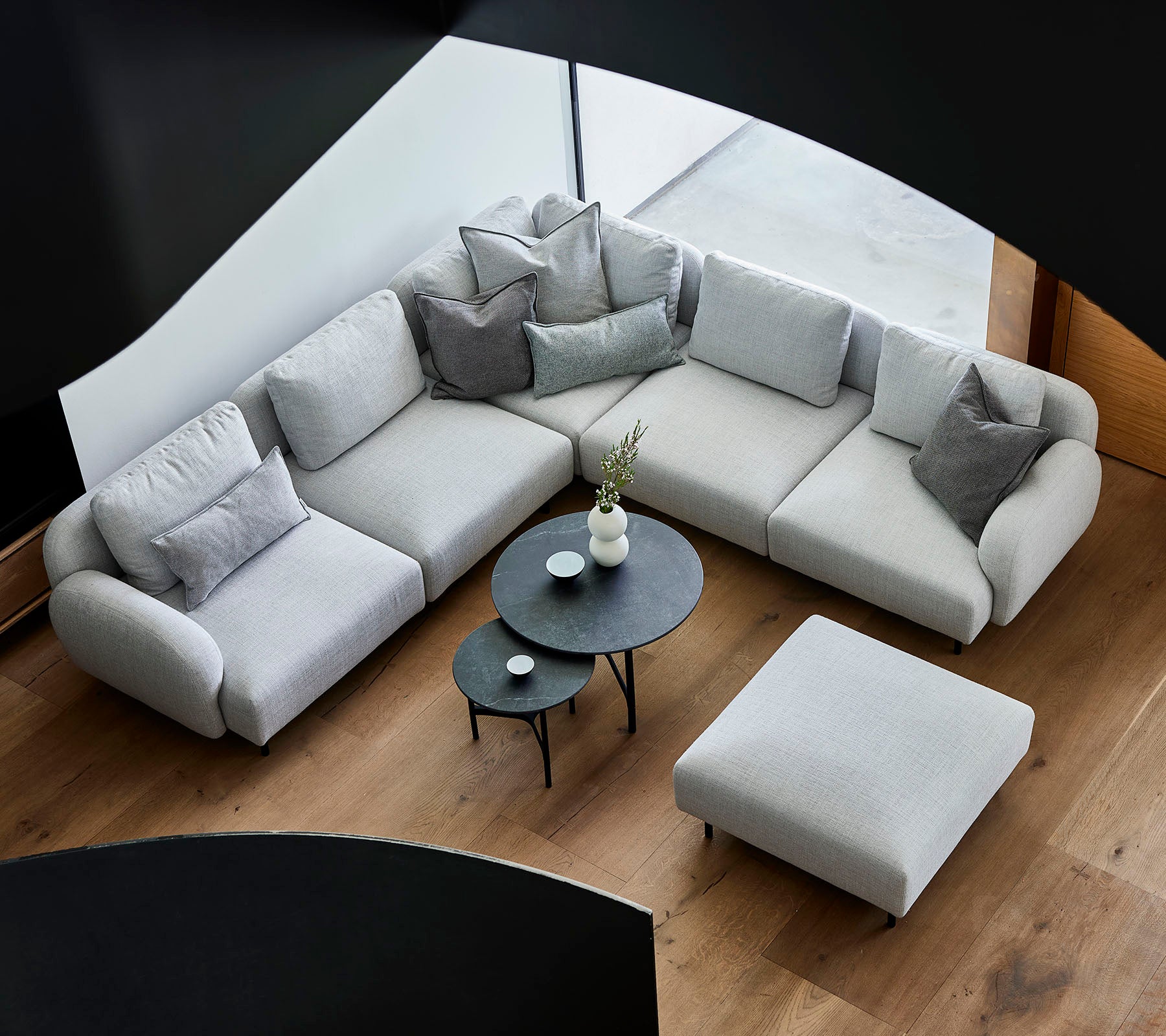 Stijlvolle grijze hoekbank met decoratieve kussens, vergezeld van een elegant rond salontafel en een ottoman op een houten vloer.