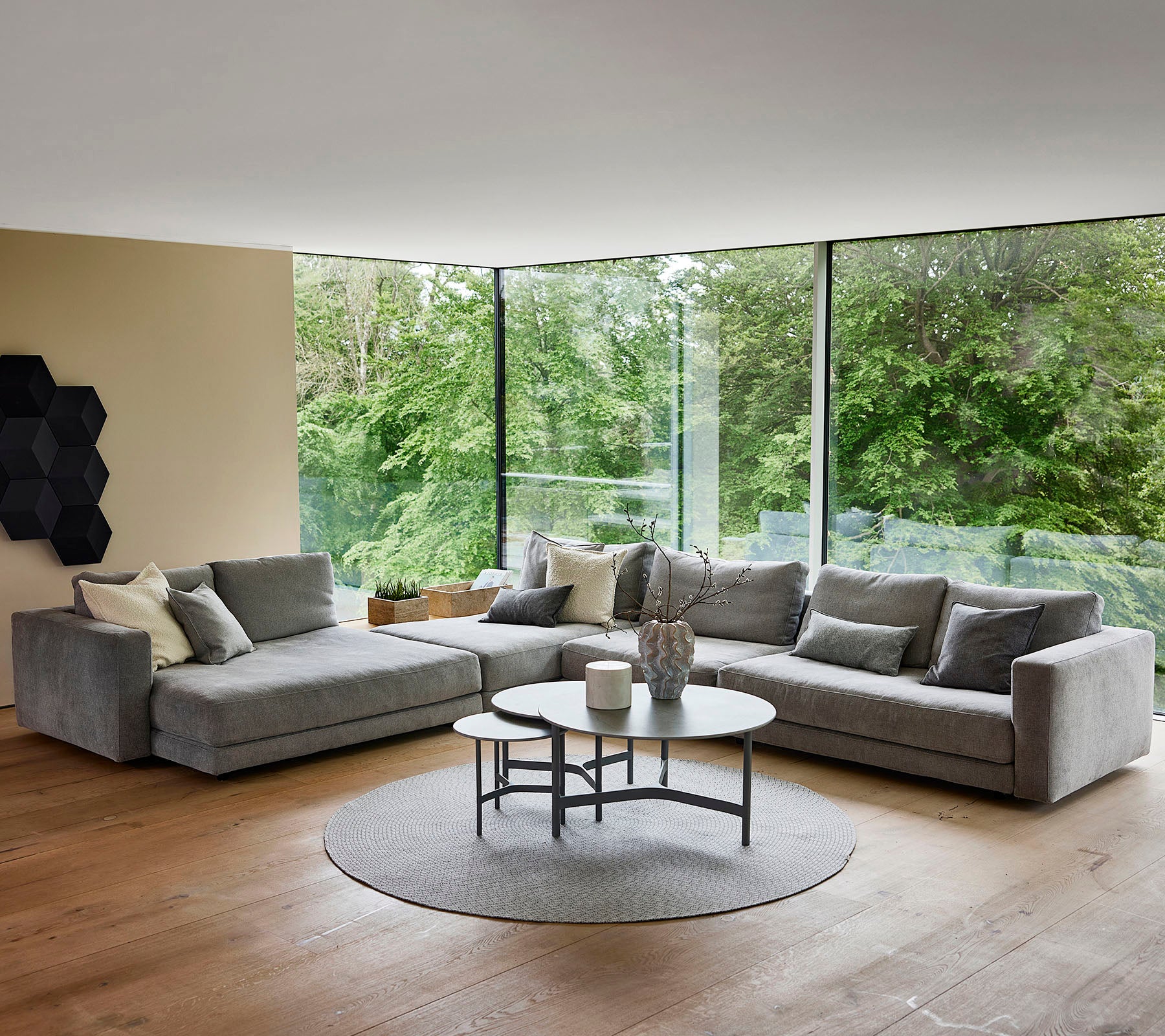 Een moderne woonkamer met een grijze hoekbank met decoratieve kussens, een ronde salontafel en een cirkelvormig tapijt, omringd door groen.