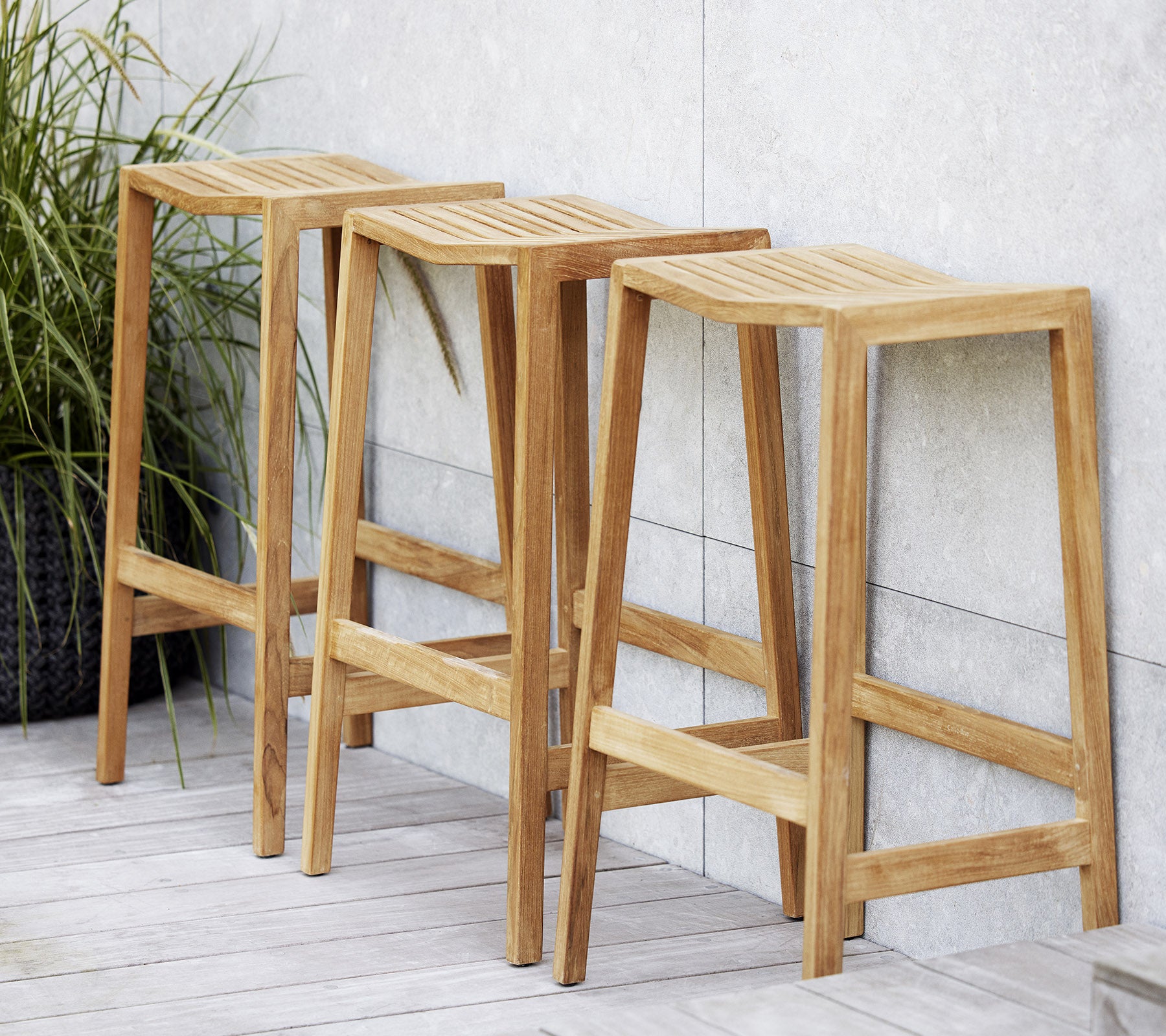 Drie houten flip-barstoelen met gesleufde zittingen, met een minimalistisch ontwerp en strakke lijnen, gepositioneerd naast een decoratieve plant.