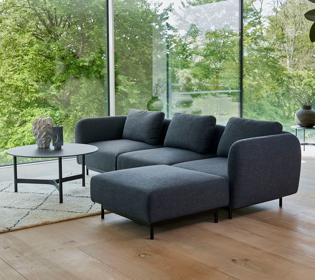 een moderne grijze hoekbank met pluche kussens, gecombineerd met een sleek rond salontafel op een licht tapijt, omringd door groen.
