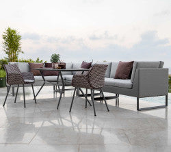 Moderne buitenzitarrangement met een grijze hoekbank, stijlvolle stoelen en een glazen tafel, versierd met decoratieve kussens.