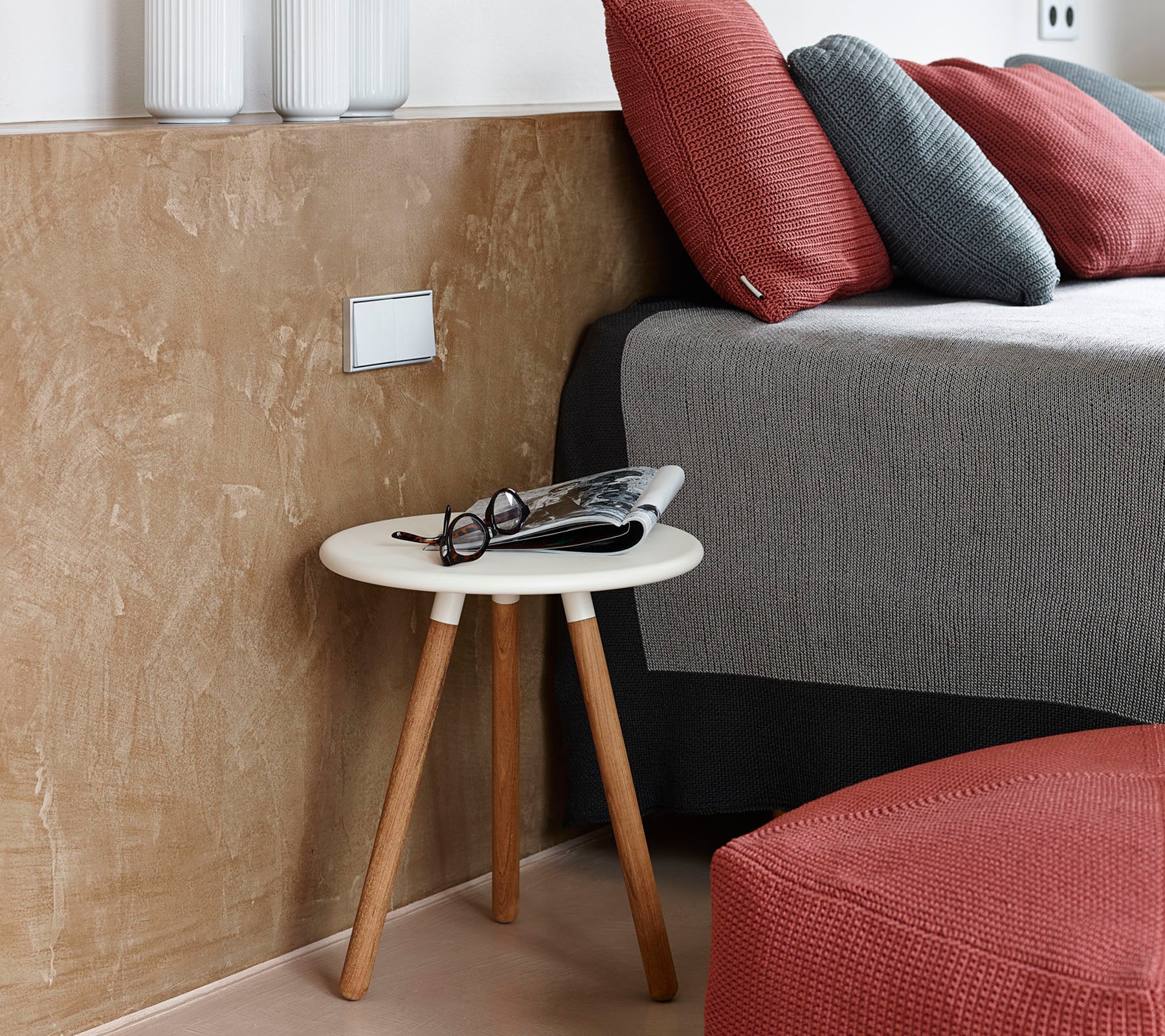 Een gezellige slaapkamer scène met een laag bijzettafeltje met brillen en een tijdschrift naast een net opgemaakt bed met gestructureerde kussens.