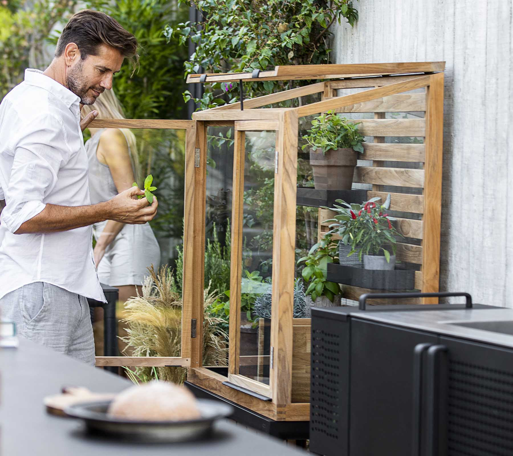 Man die planten verzorgt in een glazen display-eenheid, omringd door groen, met een moderne keukeninrichting op de voorgrond.
