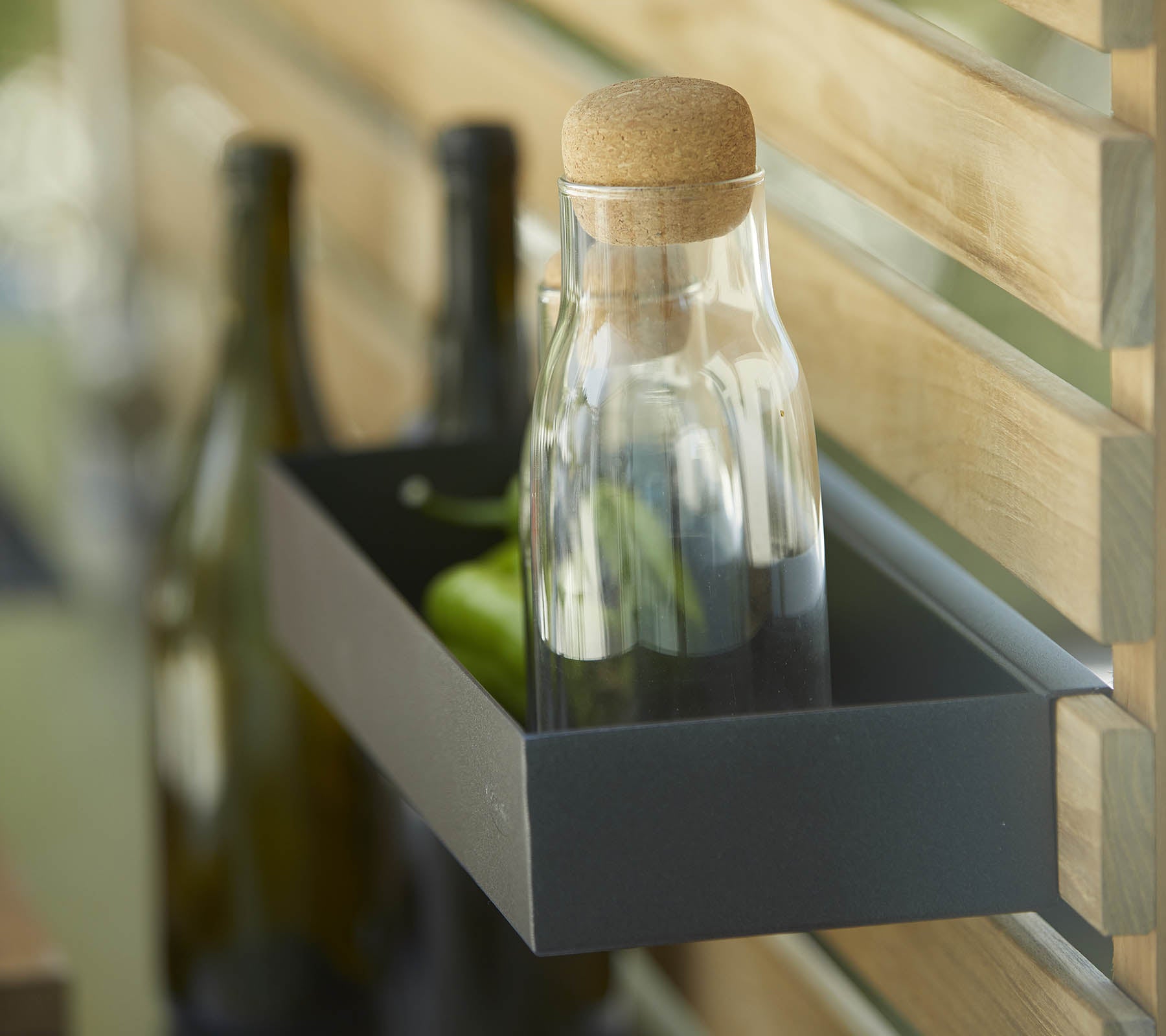 Een glazen fles met een kurk staat op een plank naast verschillende donkere flessen, tegen een houten achtergrond.