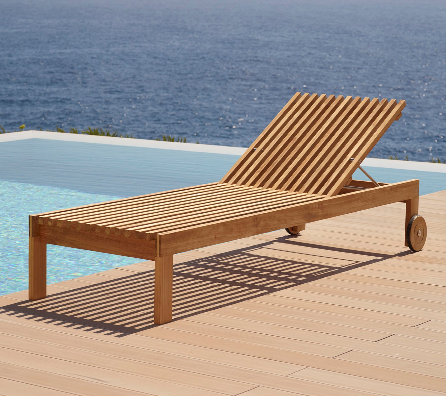 Houten loungestoel met lamellenontwerp, voorzien van wielen en geplaatst naast een zwembad met uitzicht op het water op de achtergrond.