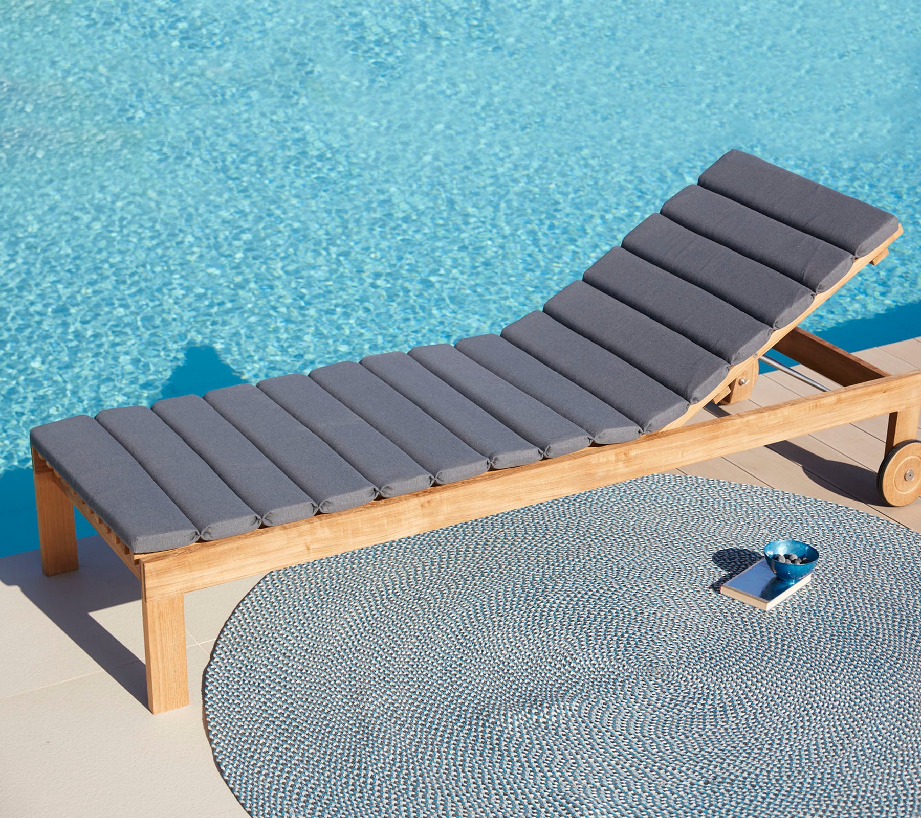 Comfortabele grijze loungechair naast een ronde mat en een kleine tray, met een rustige blauwe wateroppervlak op de achtergrond.