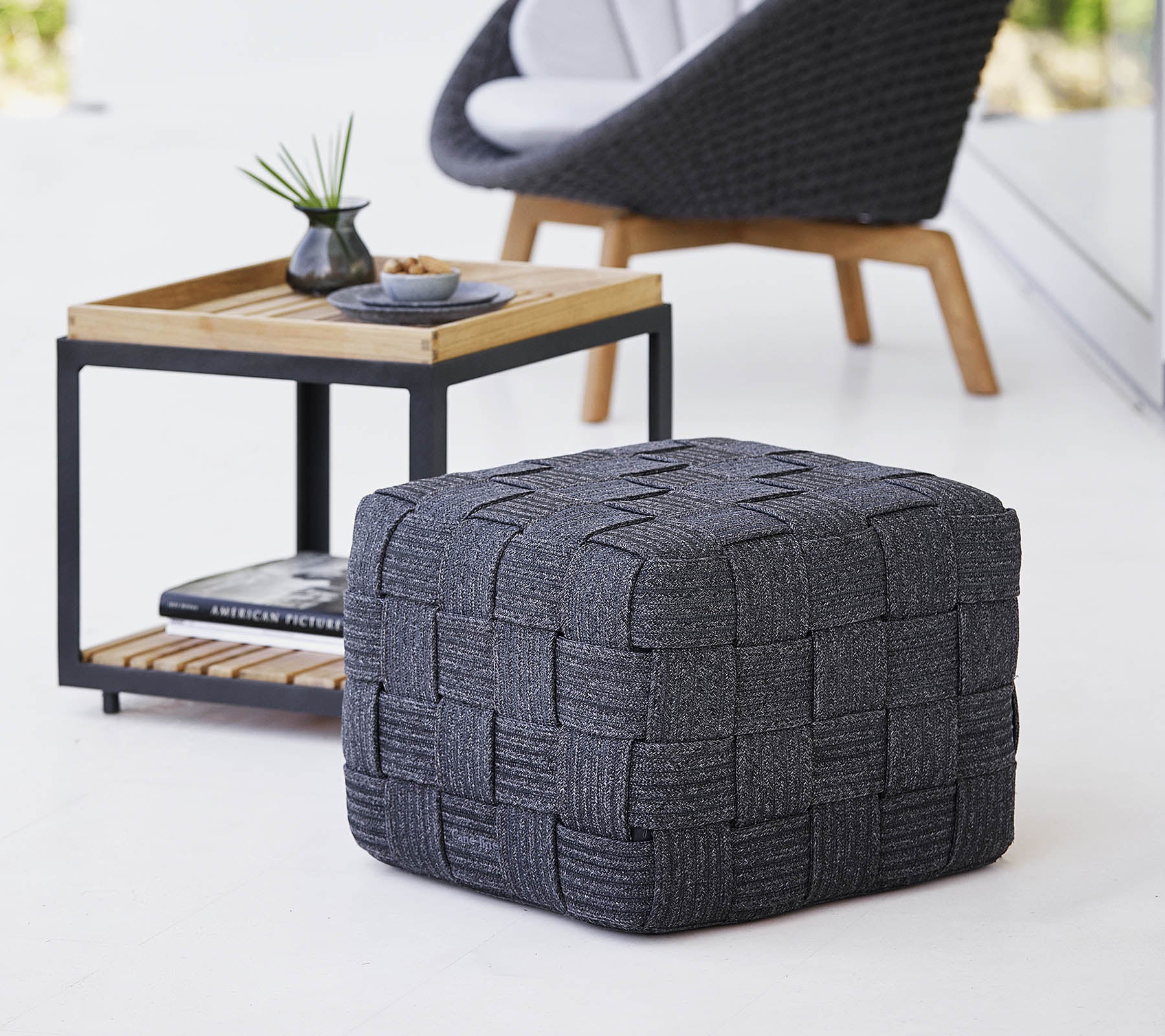 Een gestructureerde grijze geweven poef geplaatst bij een houten bijzettafel met een minimalistisch ontwerp, met kleine decoratieve items.