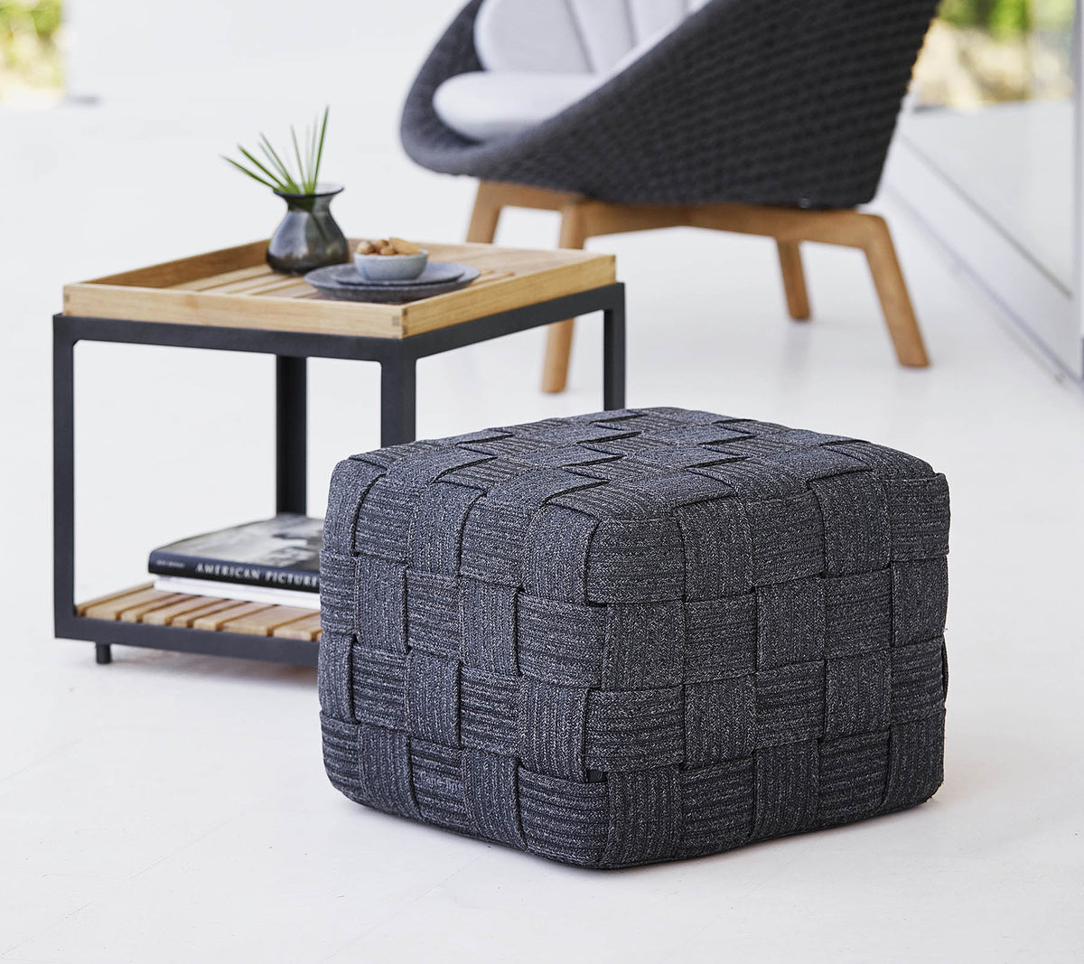 Een gestructureerde grijze geweven poef geplaatst bij een houten bijzettafel met een minimalistisch ontwerp, met kleine decoratieve items.