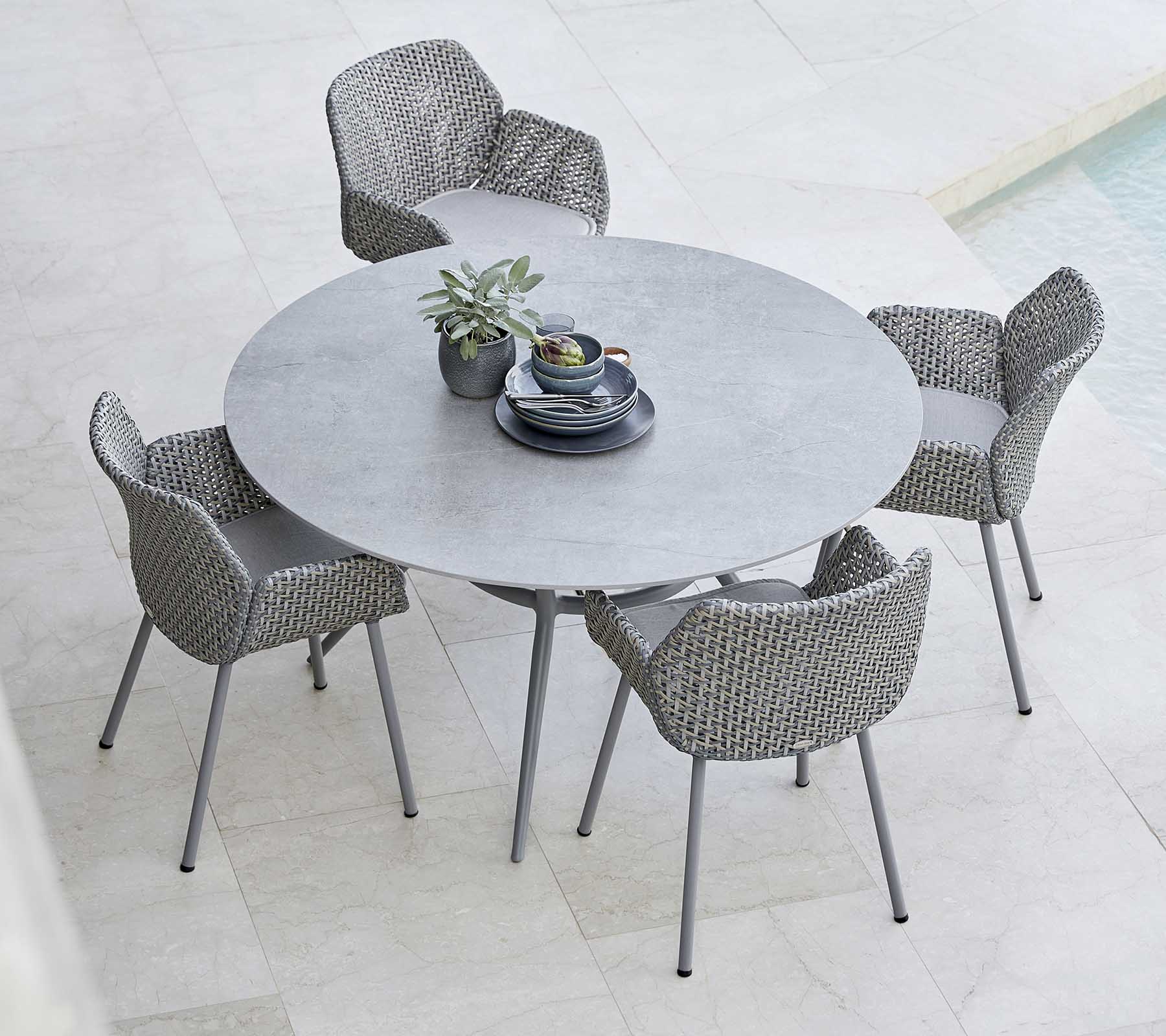 Een stijlvolle ronde tafel in combinatie met gevlochten stoelen, met een plant en serviesgoed in het midden, perfect voor buiten dineren.