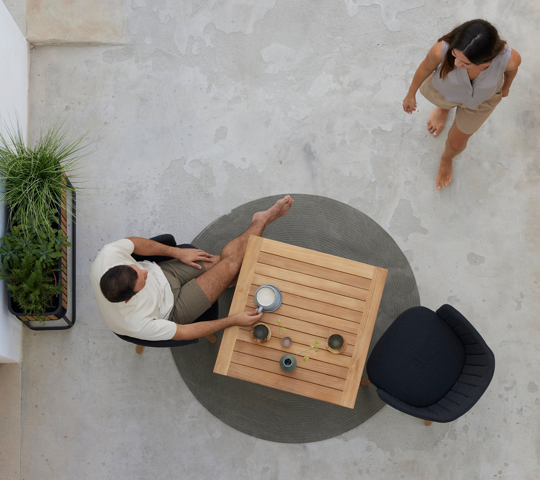 Twee mensen genieten van koffie aan een teak tafel omringd door groen, met een zwarte stoel in de buurt. Een ontspannen café sfeer wordt weergegeven.