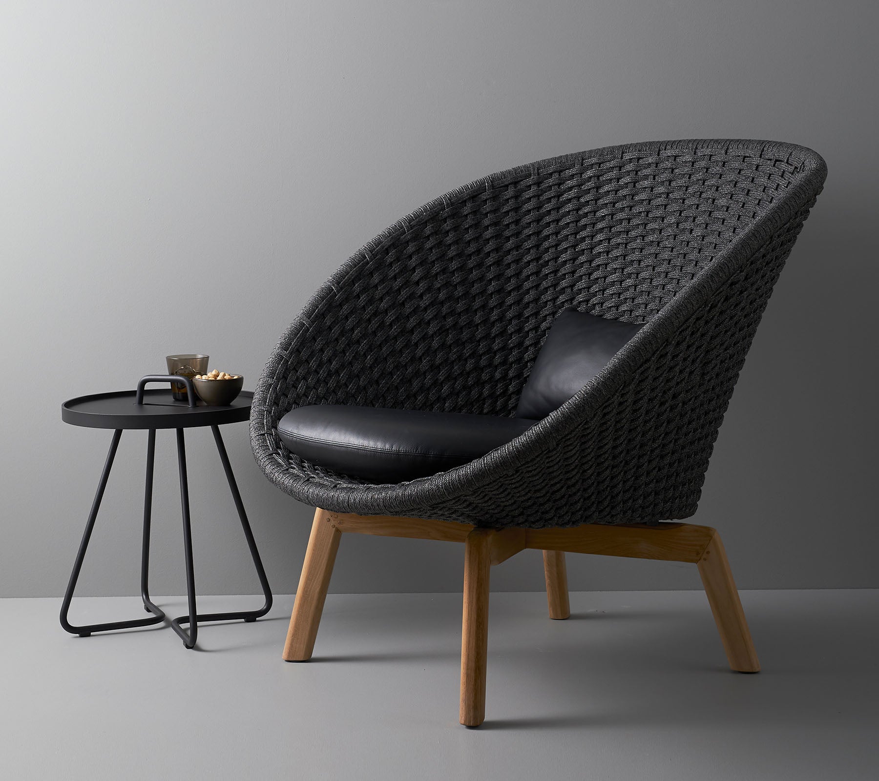 Stijlvolle gevlochten stoel met een zwart kussen en een moderne ronde salontafel, ideaal voor hedendaagse woonruimtes.