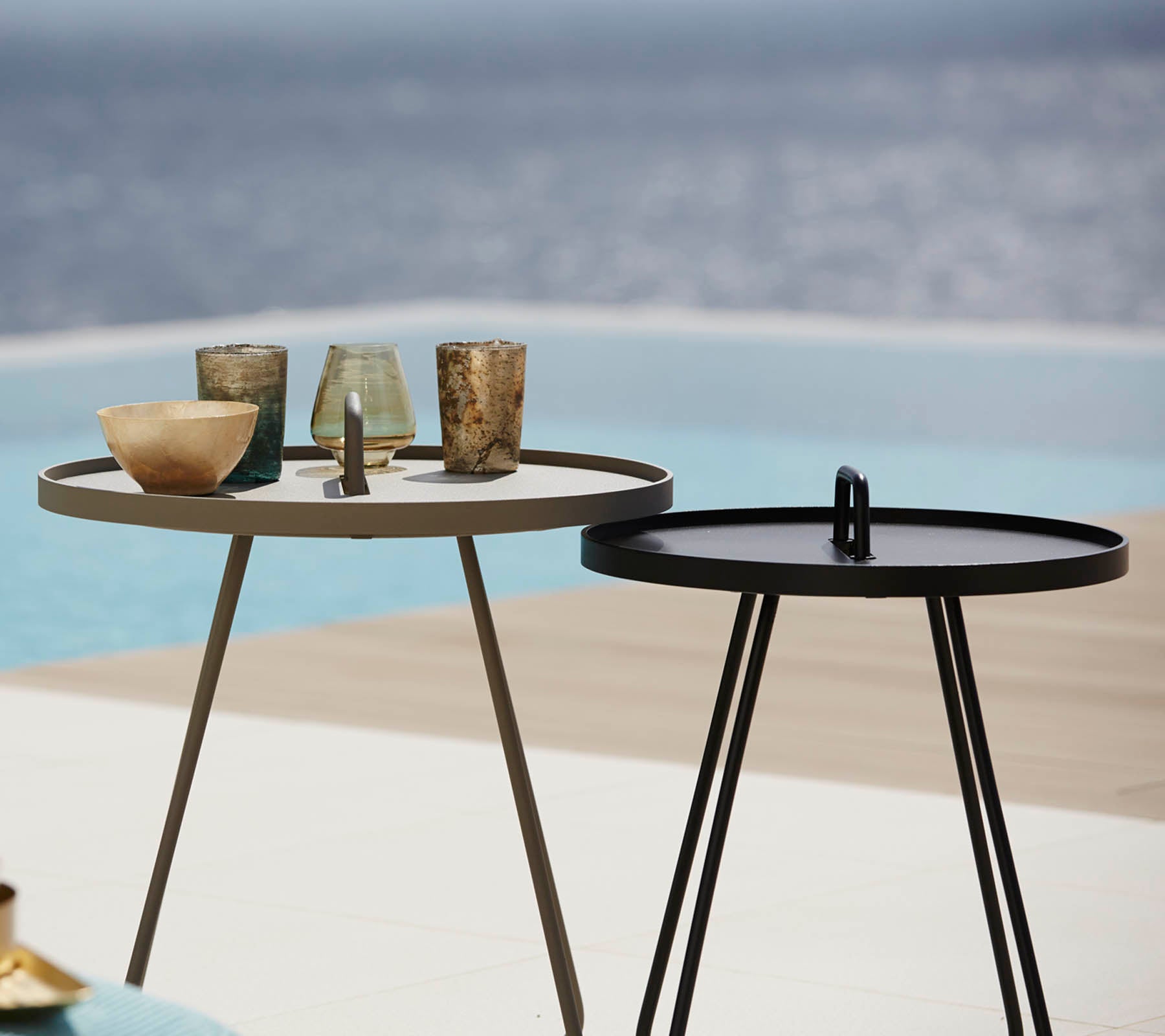 Two stylish side tables with various decorative glassware on top, placed near a serene water feature in the background.