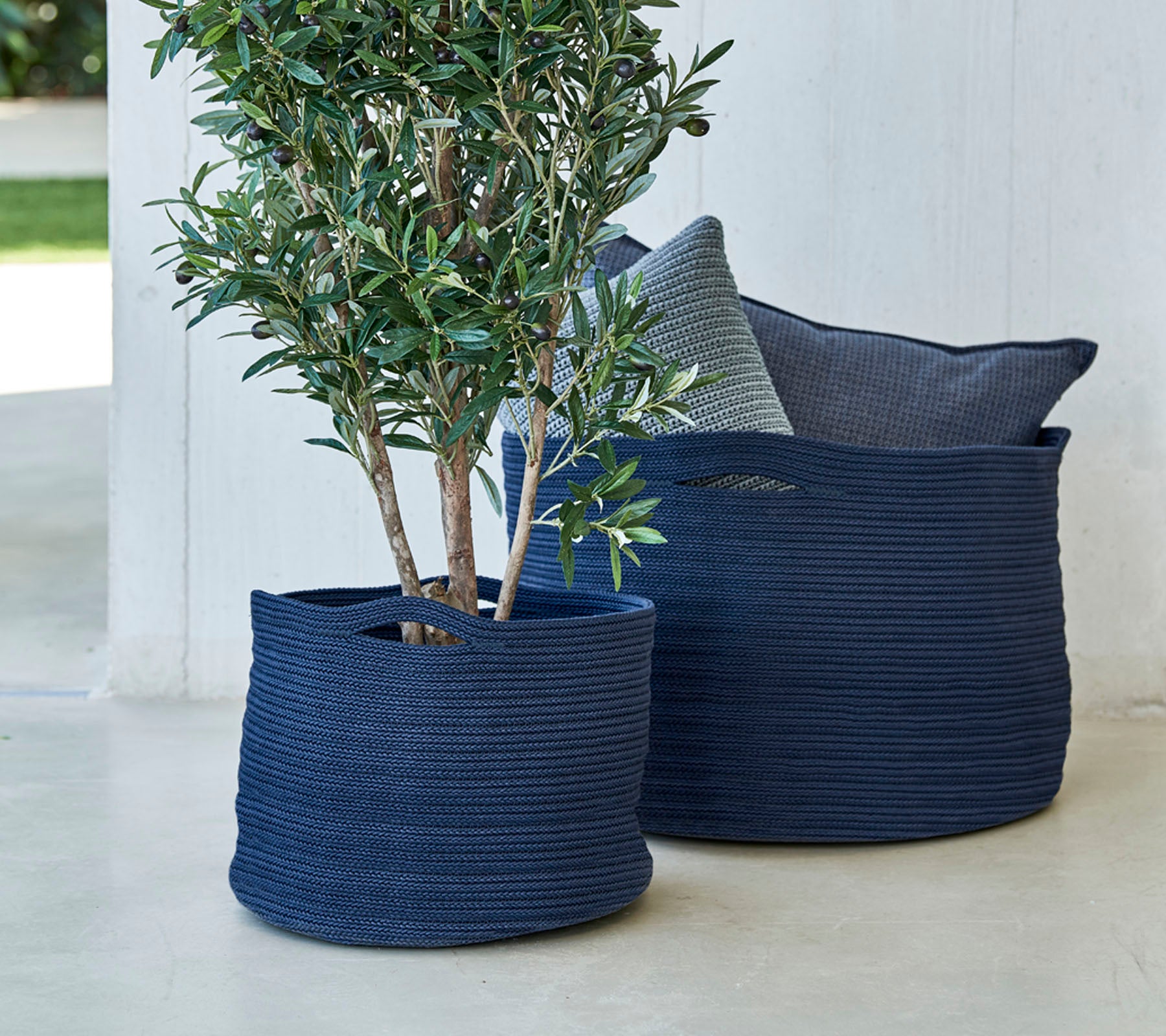 Twee marineblauwe geweven plantenbakken, waarvan er één een kleine boom bevat, vergezeld van een decoratief kussen.